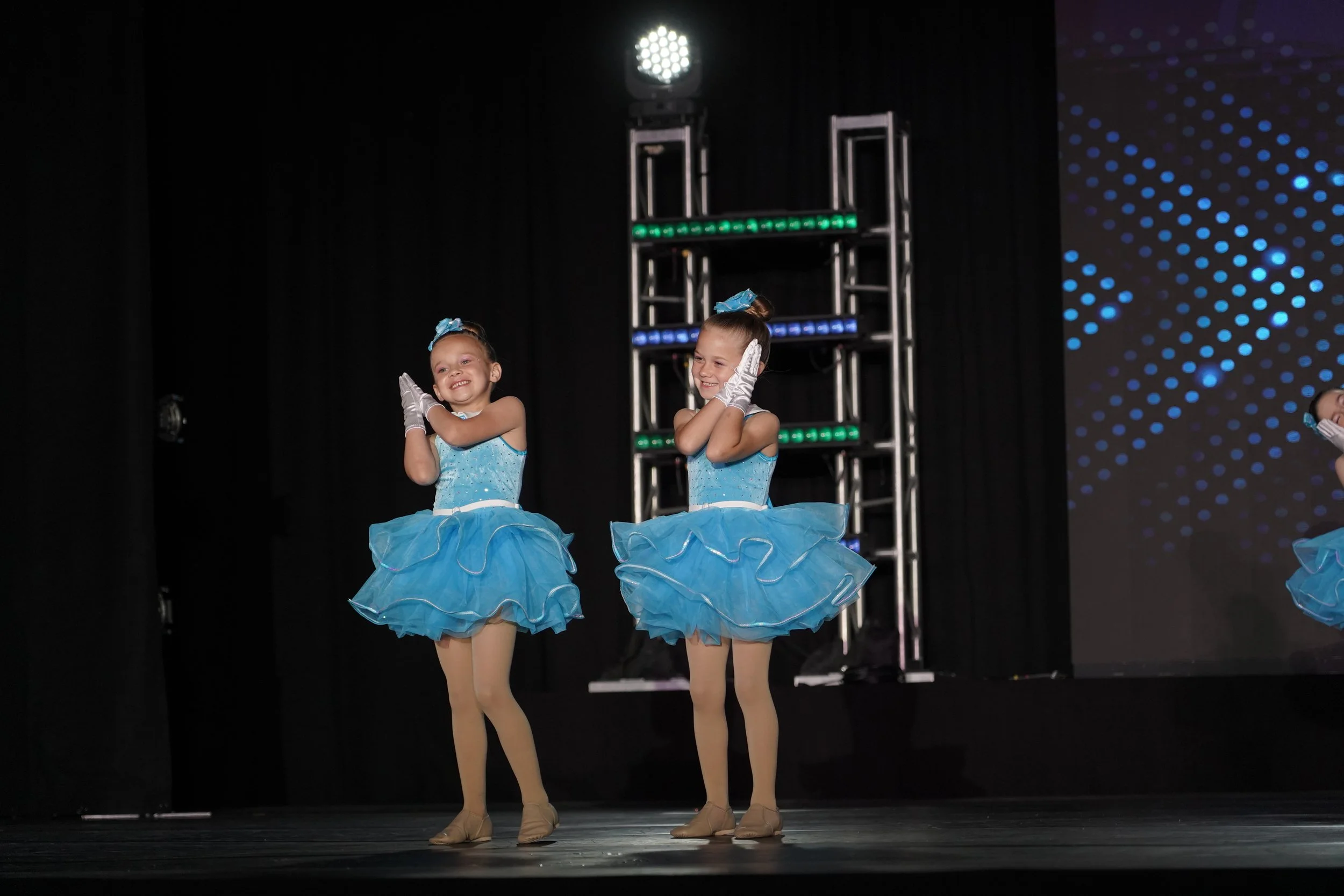Two young girls in blue costumes performing a dance on stage, with black curtains, stage lighting, and a digital backdrop.