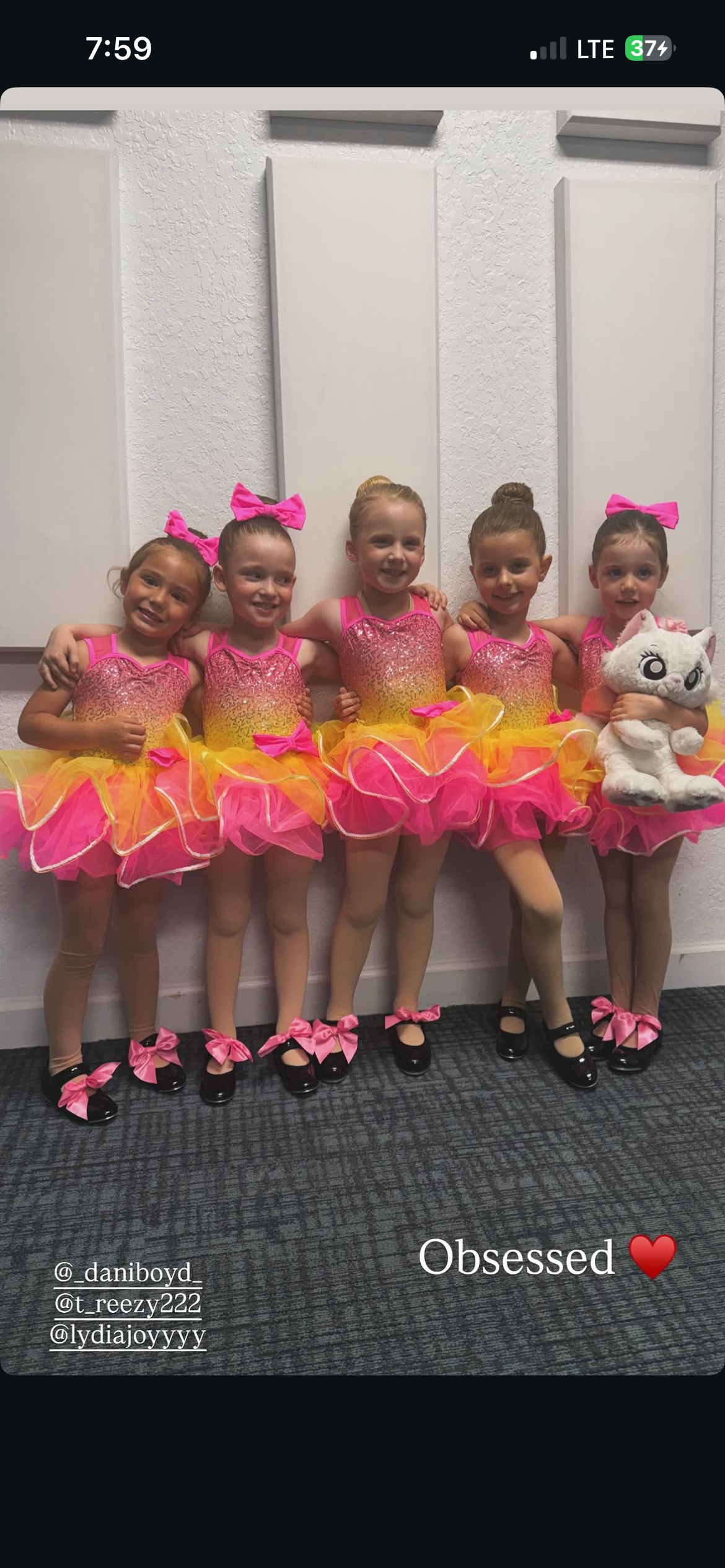 Five young girls dressed in pink and yellow ballet costumes, standing together with their arms around each other, smiling, holding a stuffed cat toy. They are wearing matching shoes with pink bows, and appear to be backstage or in a dressing room.