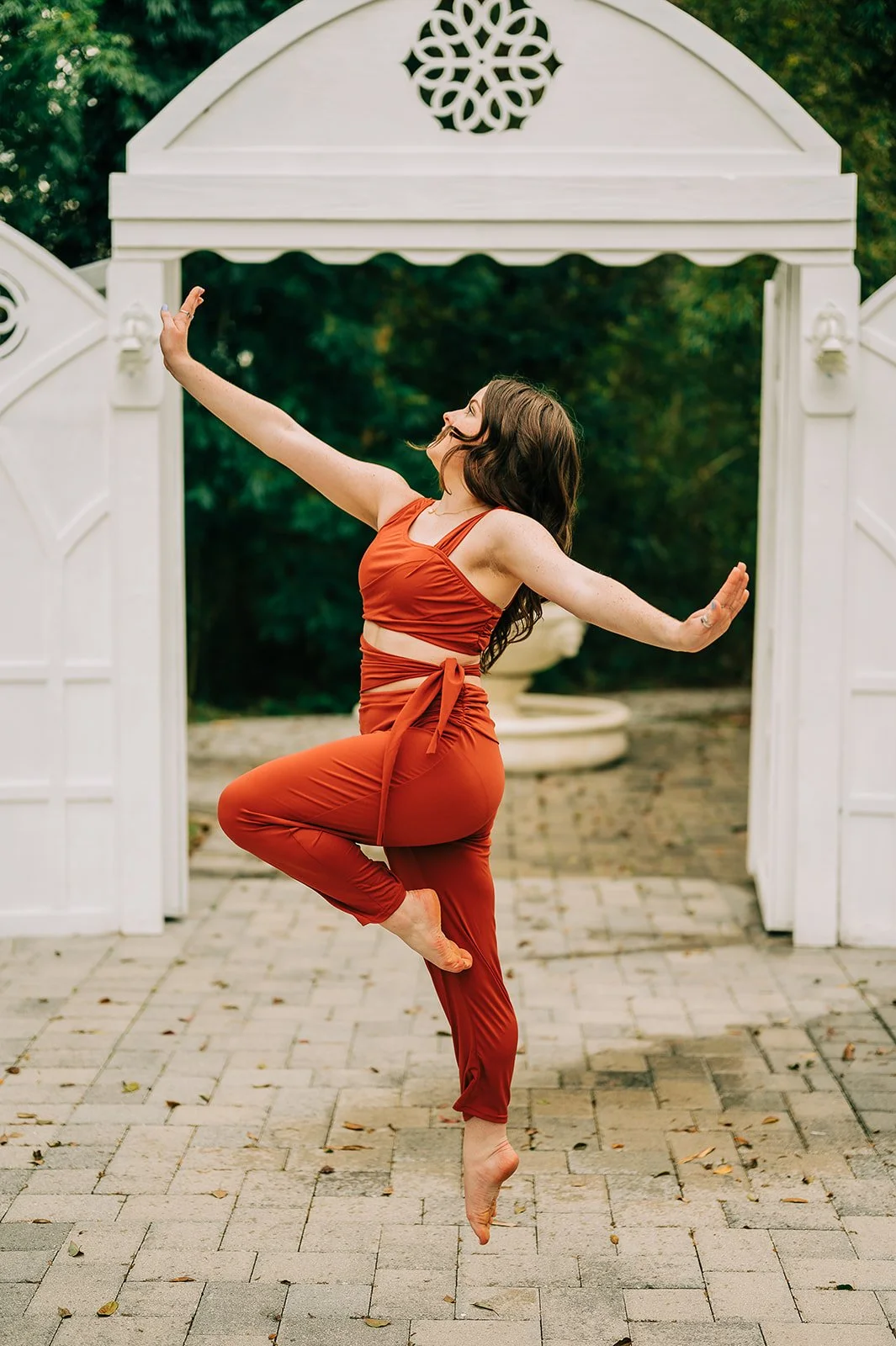 A woman in orange yoga attire practicing a yoga pose outdoors on a paved area with a white decorative archway and greenery in the background.