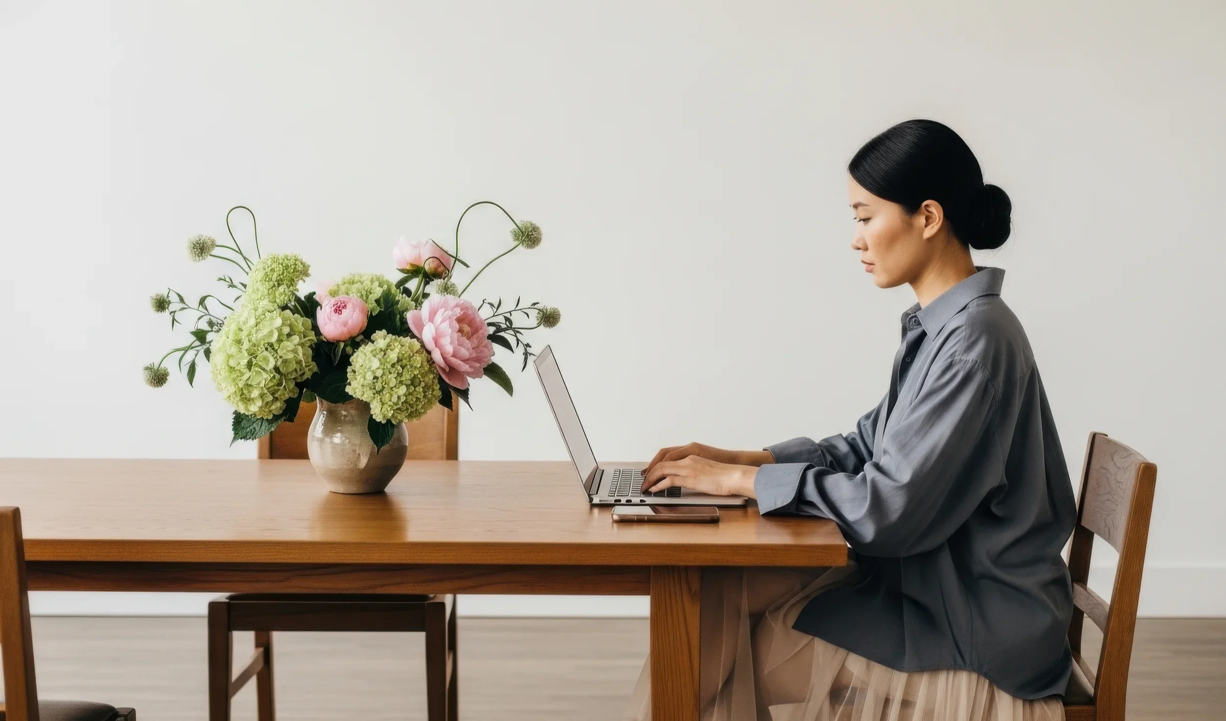 A woman sitting at a wooden table working on a laptop, with a vase of pink and green flowers next to her, against a plain white wall.