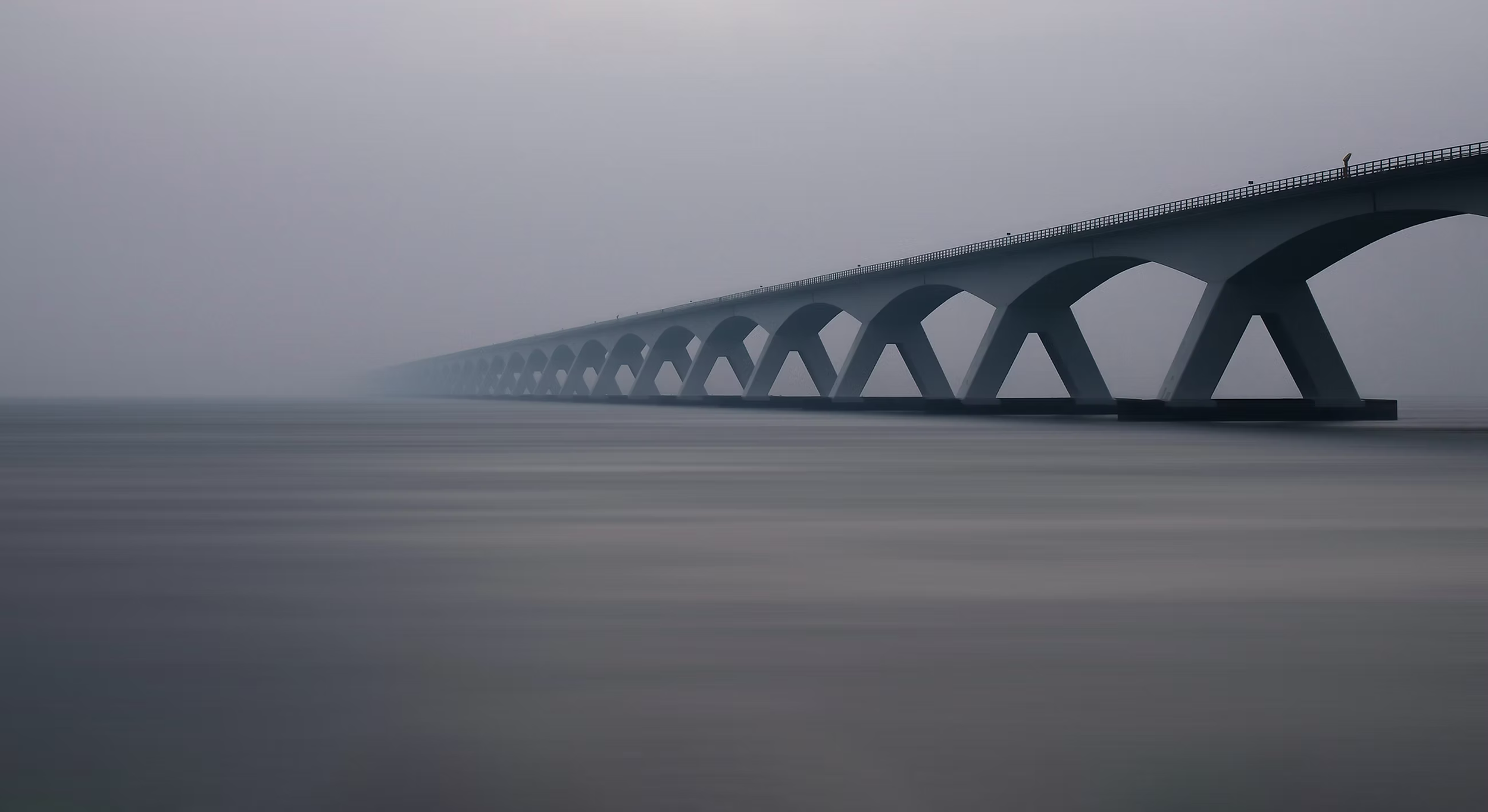 A long bridge with multiple arches extends over calm water on a foggy day.