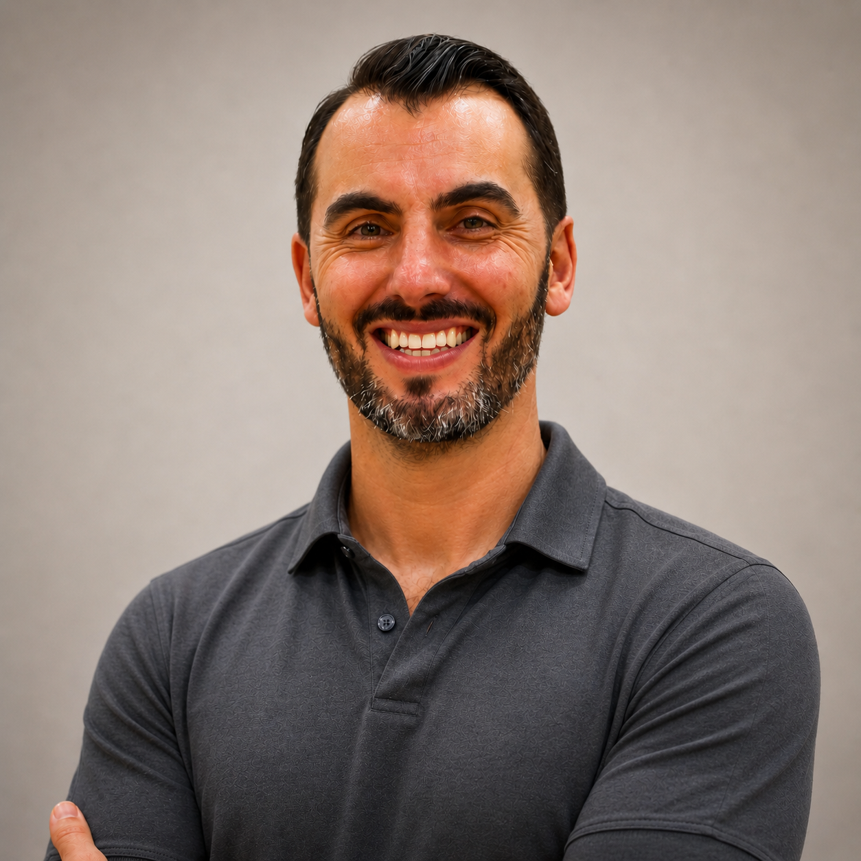 Smiling man with dark hair, beard, wearing a gray polo shirt, standing against a plain background.