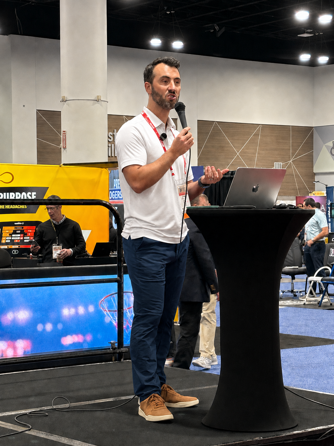 A man in a white shirt and blue pants is speaking into a microphone while standing behind a black podium at a conference. He has a laptop on the podium and is gesturing with his left hand.