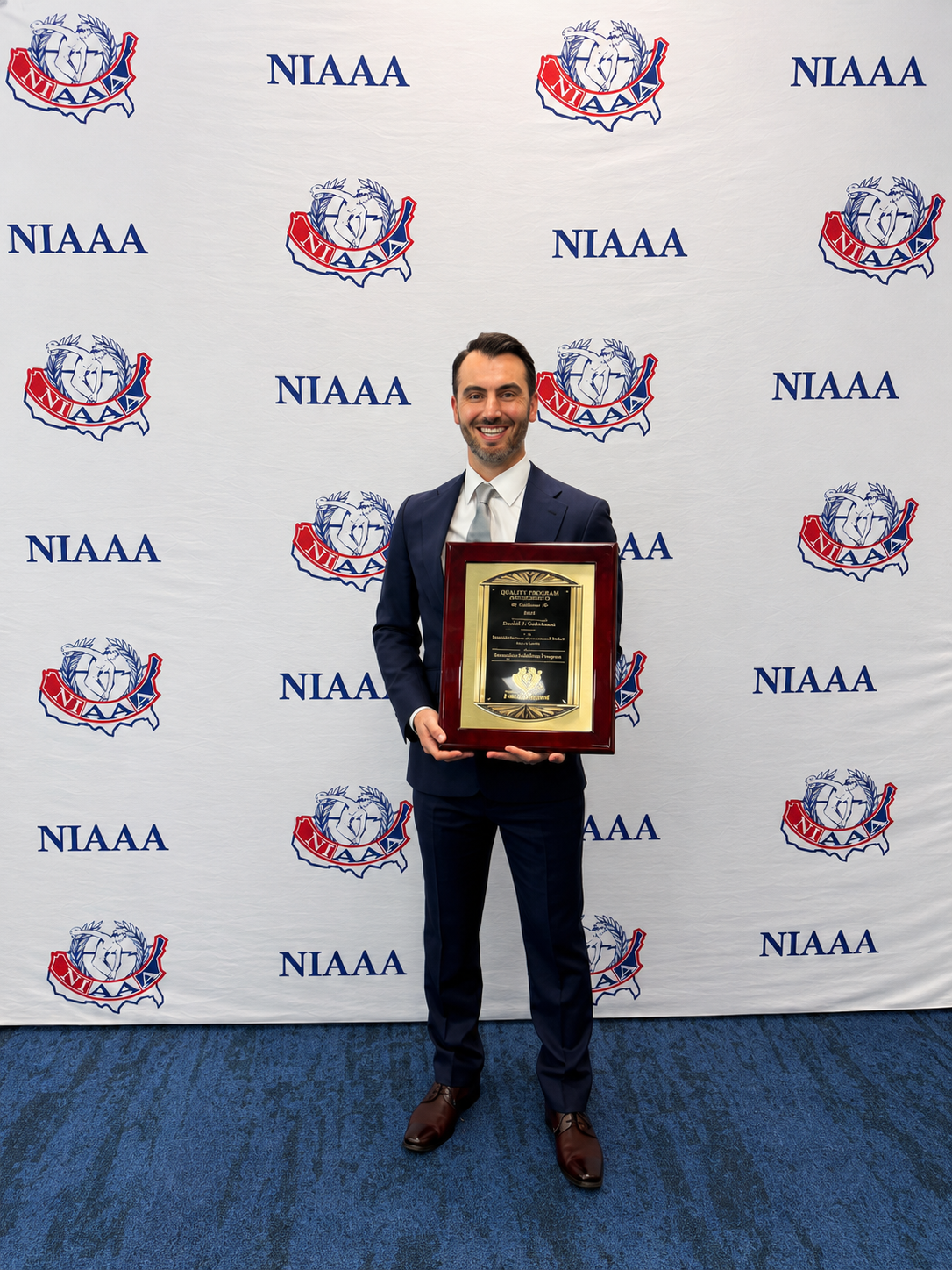 A man in a suit holding a framed award standing in front of a white backdrop with the NIAAA logo and text repeated.