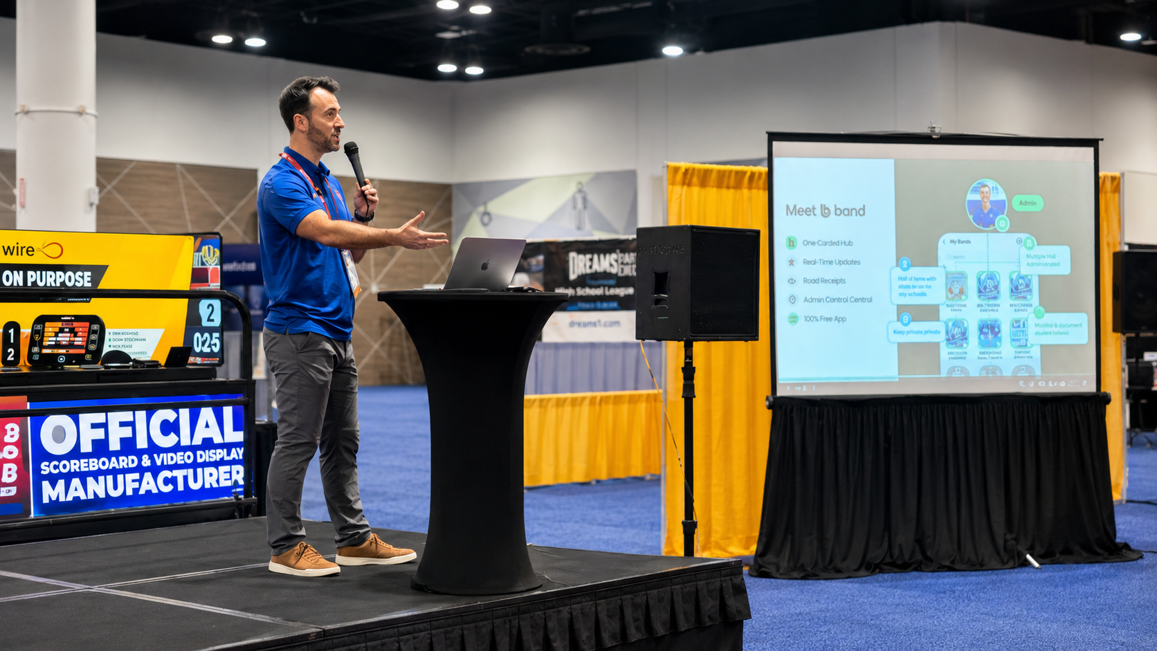 A man in a blue shirt giving a presentation at a conference, standing at a black podium with a speaker and laptop, with a large screen displaying a presentation slide behind him, in an exhibition space with banners and booths.