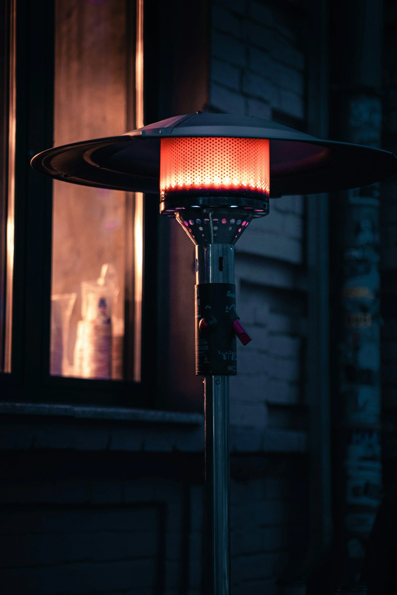 A patio heater with a black top and a glowing red heating element, standing outside near a building during evening or night.