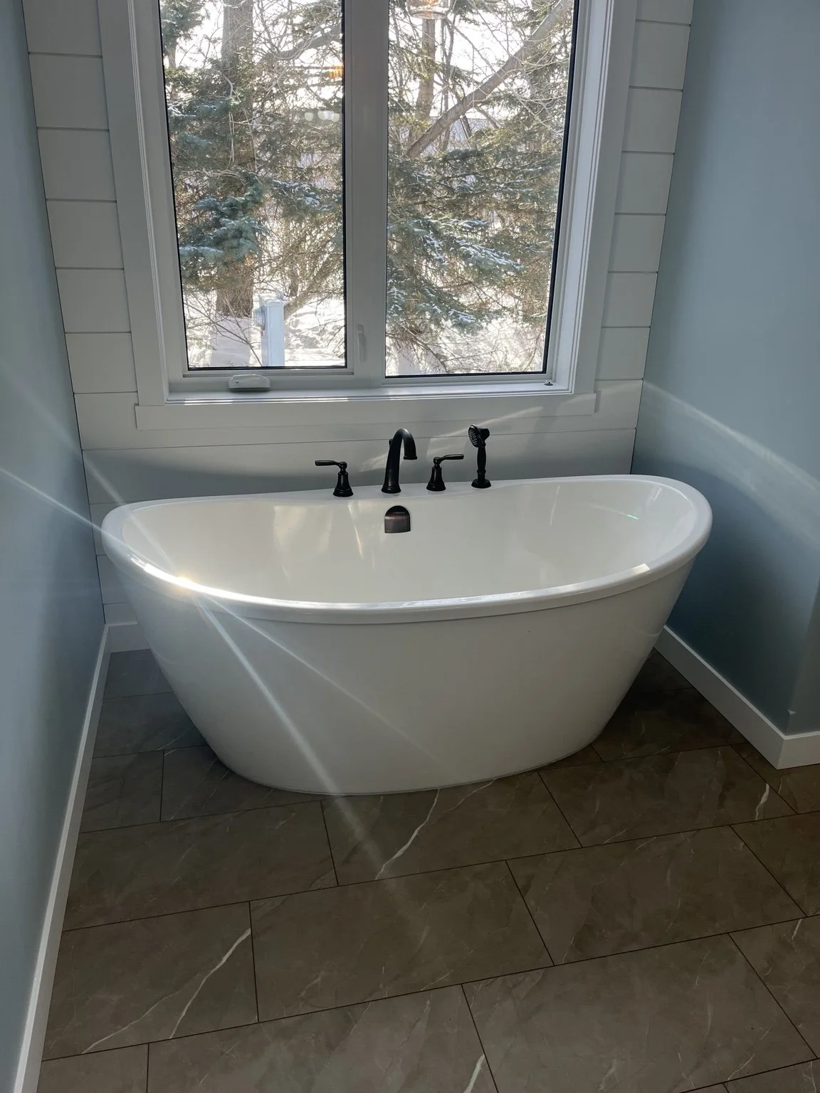 A bathroom with a white bathtub placed underneath a large window showing trees outside. The bathtub has black fixtures, including a faucet and handheld shower, on a beige tiled floor.