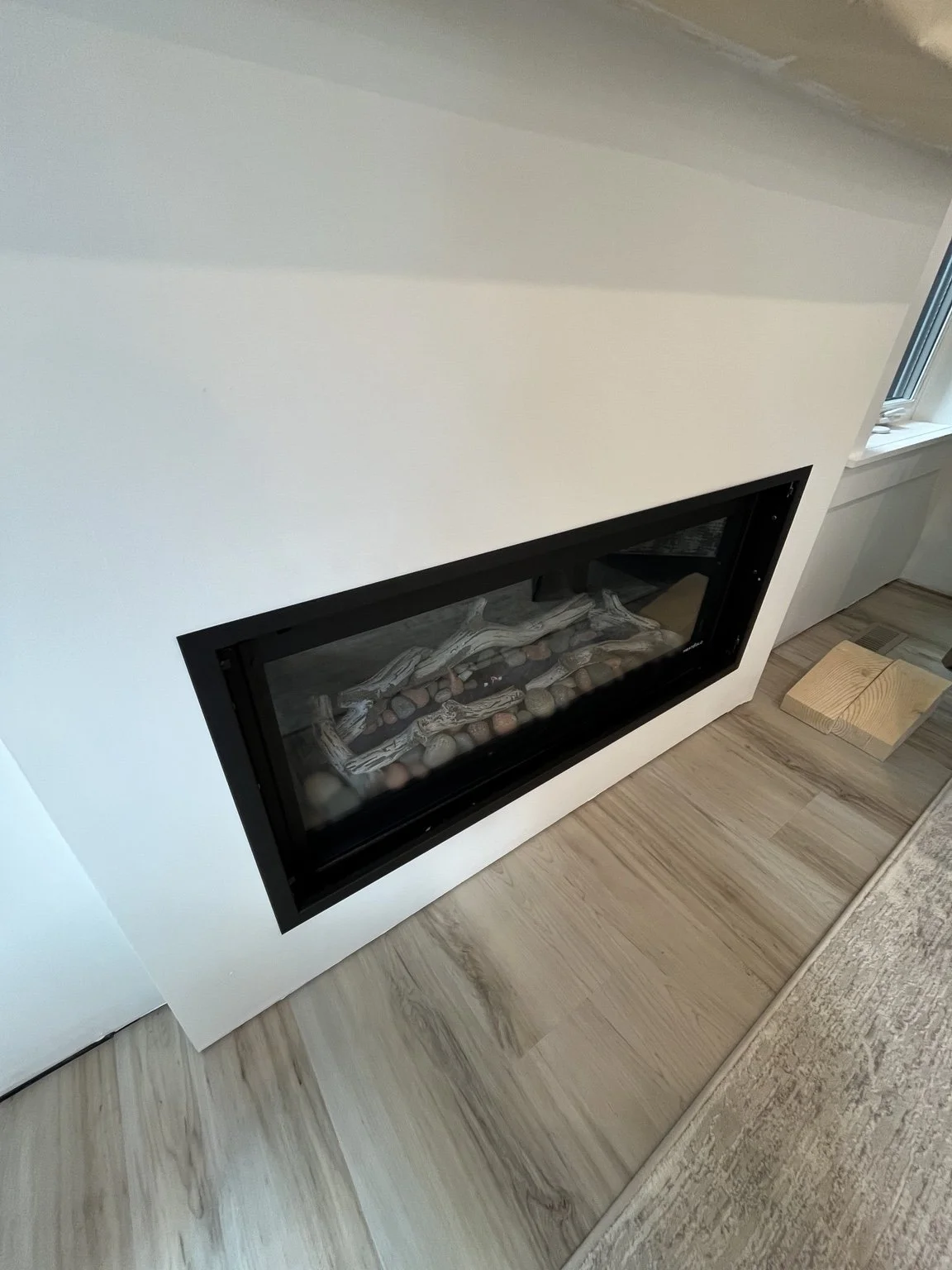 Modern indoor fireplace with black frame built into a white wall, decorated with driftwood and stones, situated on light wooden flooring.