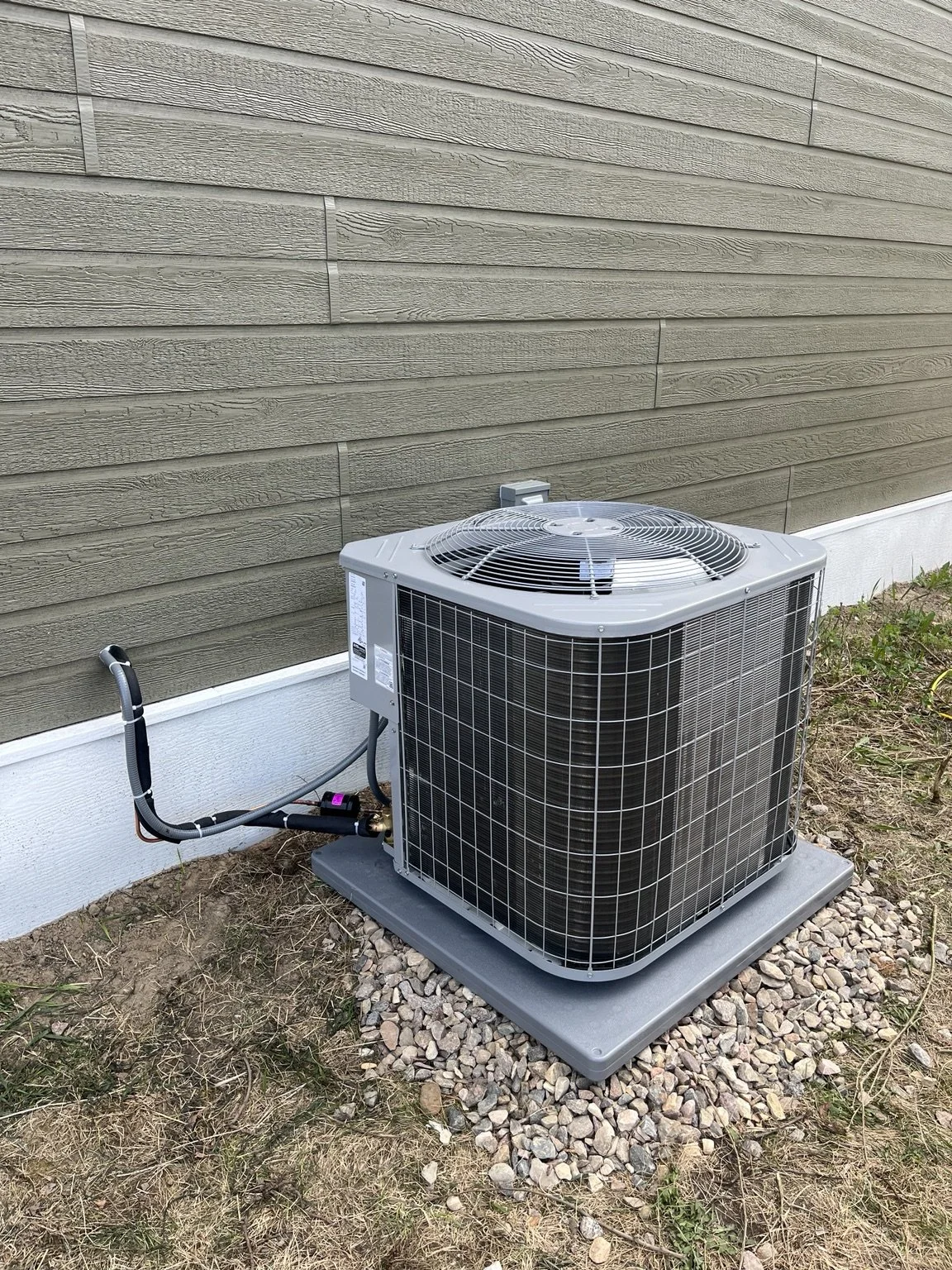 An outdoor air conditioning unit installed beside the beige house exterior, on a gravel base with grass and small plants nearby.