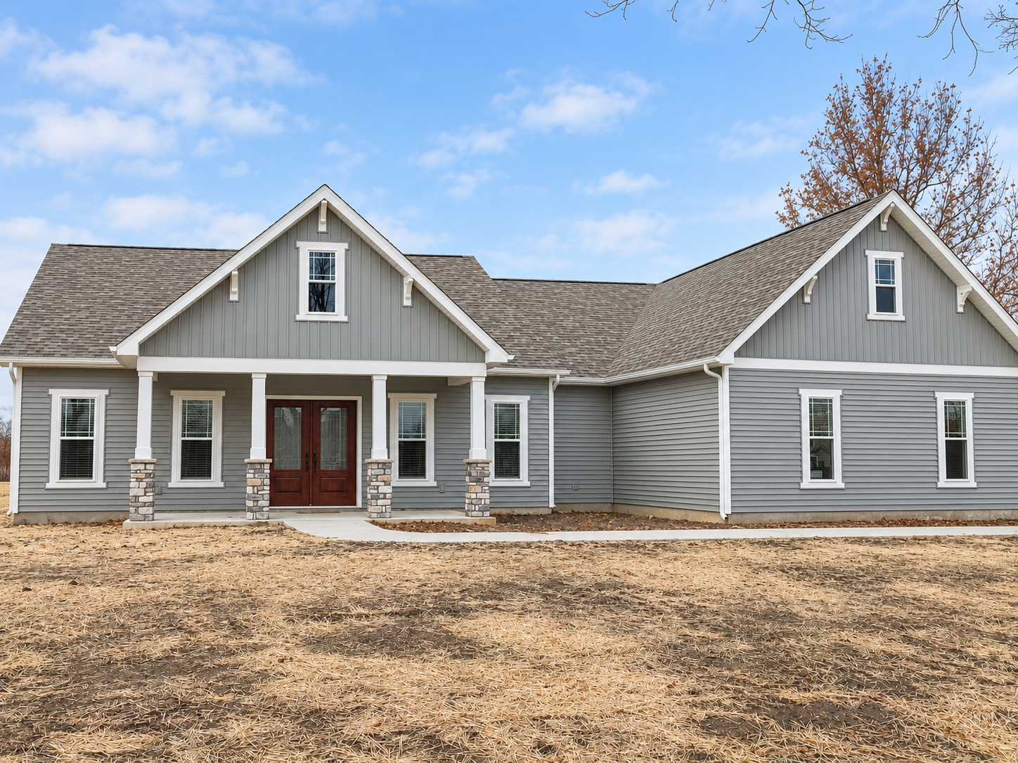 A newly built, modern house with gray siding, white trim, and a brown front door, located on a plot with dry dirt ground and a clear blue sky background.