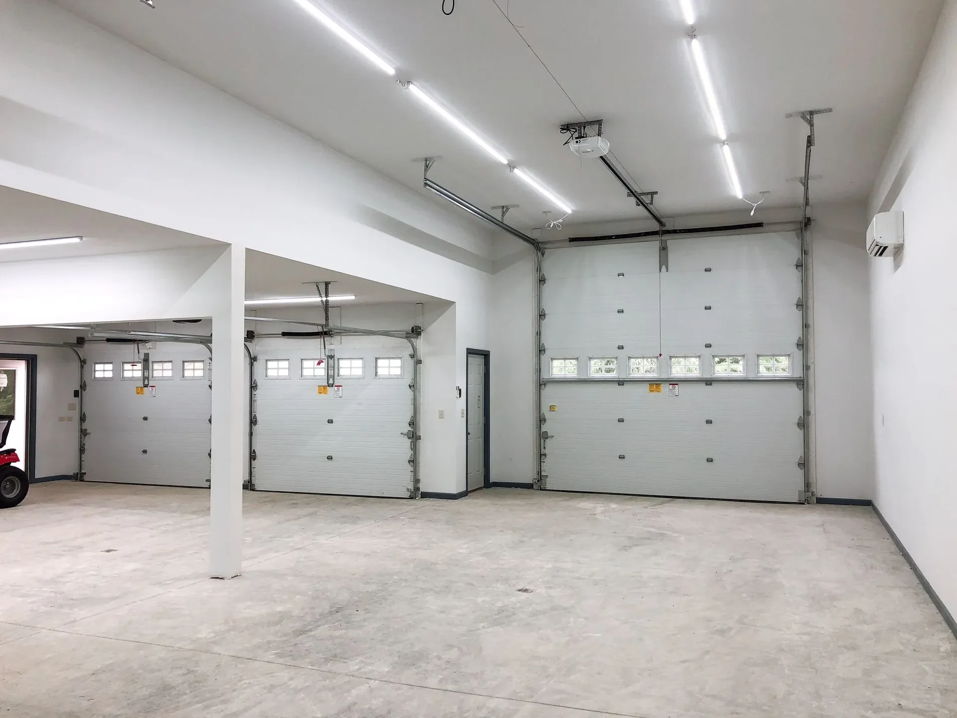 Empty garage with three closed white garage doors, fluorescent lighting, and a gray concrete floor.