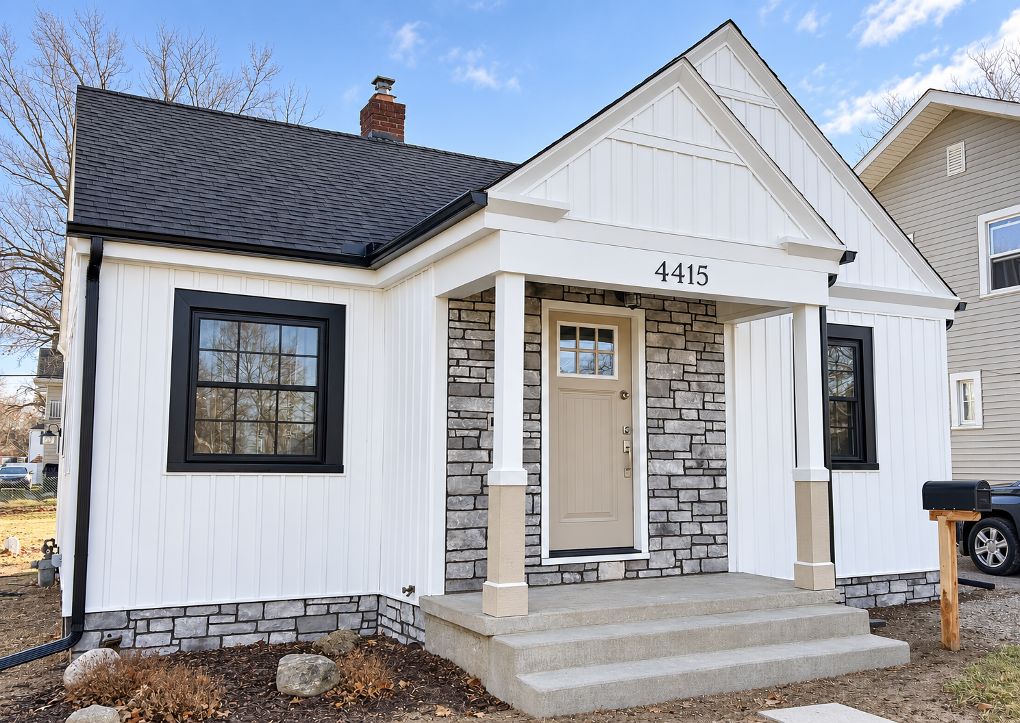 Newly built house with white vertical siding, black window frames, gray brick accents, and a gray front door with a small window. The house number 4415 is above the porch, which has concrete steps leading to the entrance. The roof is black, and there are leafless trees in the background under a partly cloudy sky.