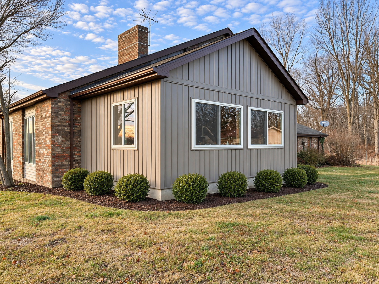 A house with a brick chimney, gray siding, white-framed windows, a landscaped yard with small bushes, a grassy lawn, and trees in the background, under a blue sky with scattered clouds.