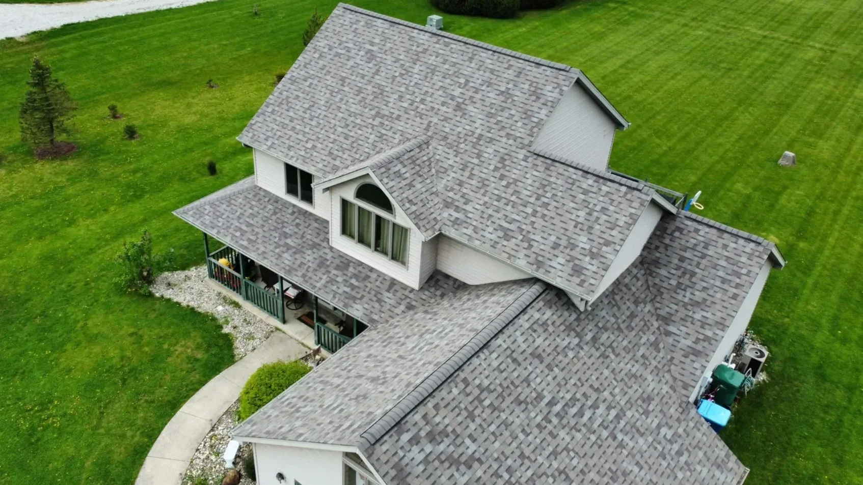 An aerial view of a two-story house with gray shingle roof, white siding, and a covered front porch surrounded by green lawn and trees.