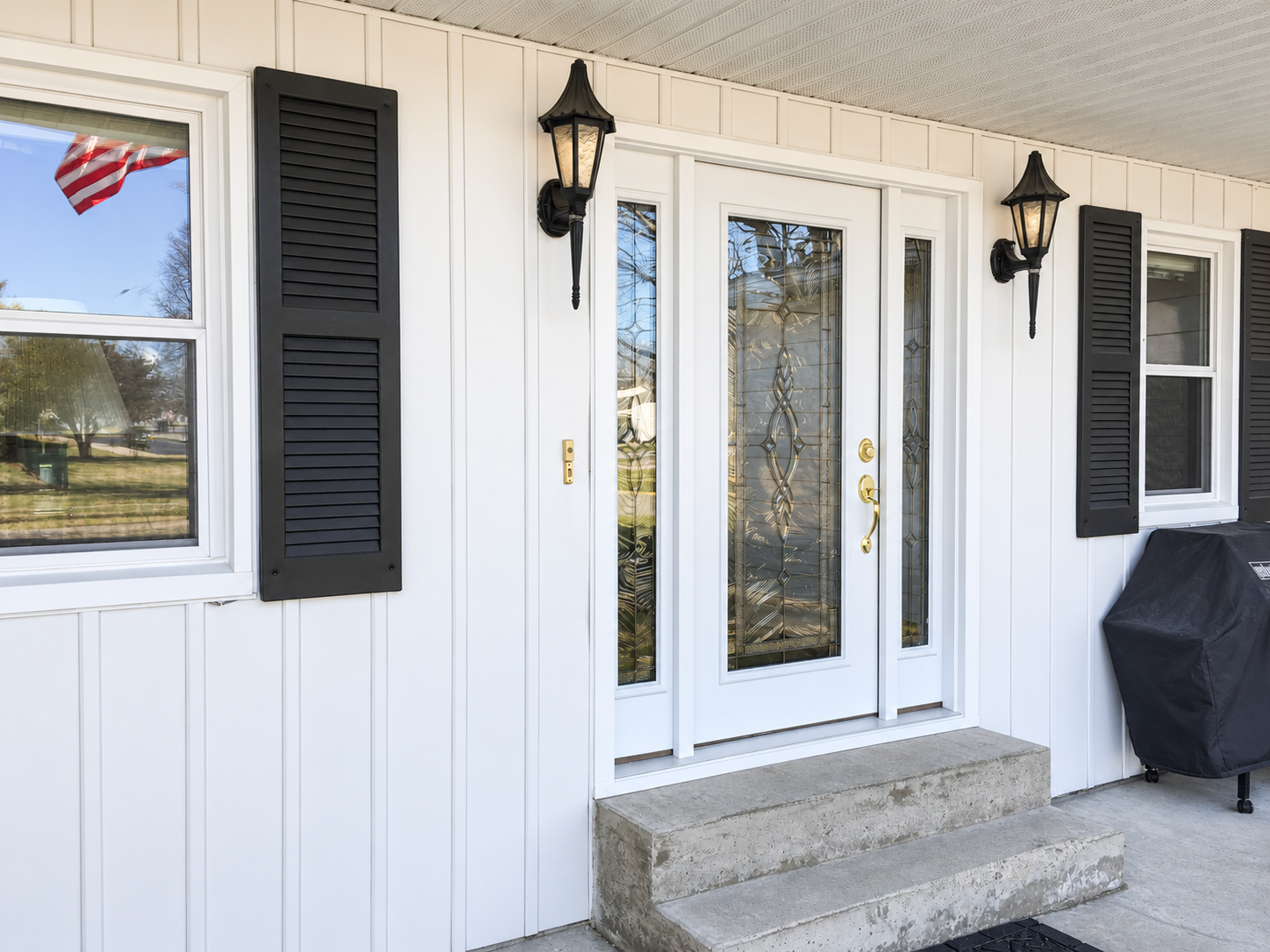 White front door with decorative glass panels and gold hardware, flanked by two black wall-mounted lanterns, with black shutters on adjacent windows, a concrete step at the entrance, and a covered porch with a hanging ceiling.