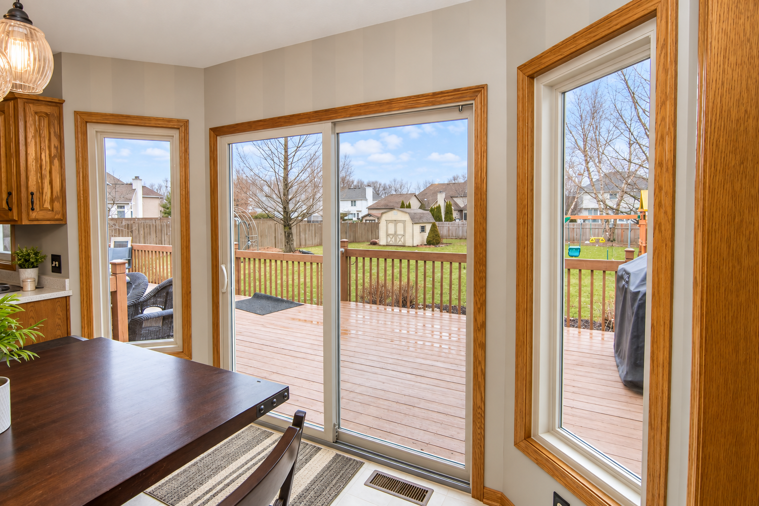 Bright kitchen with large sliding glass door and window overlooking a backyard with a deck, trees, a shed, a trampoline, and houses in the distance.