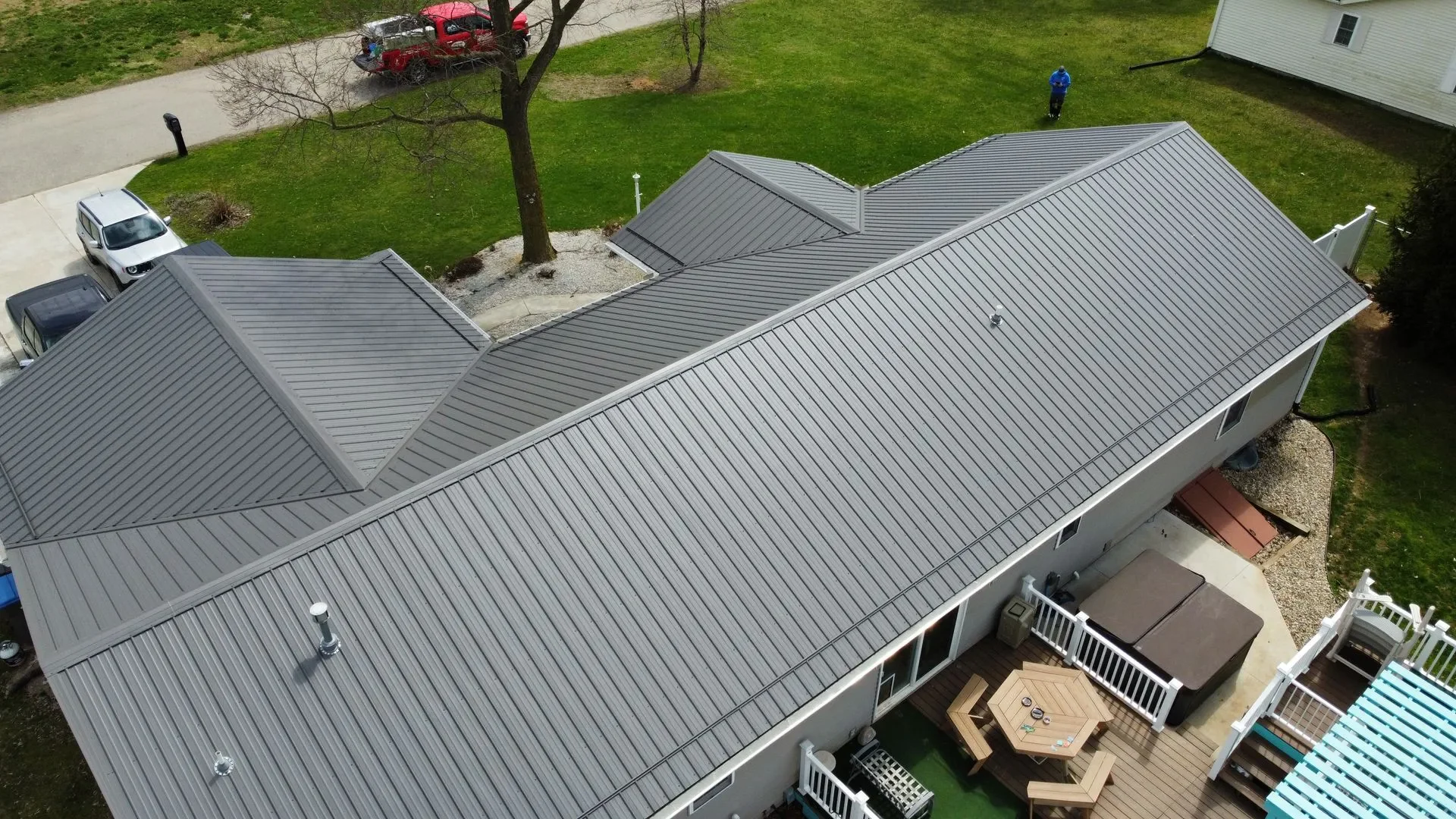 Aerial view of a house with a gray metal roof, a backyard patio with wooden furniture, and people on a lawn, with a parking area and trees nearby.