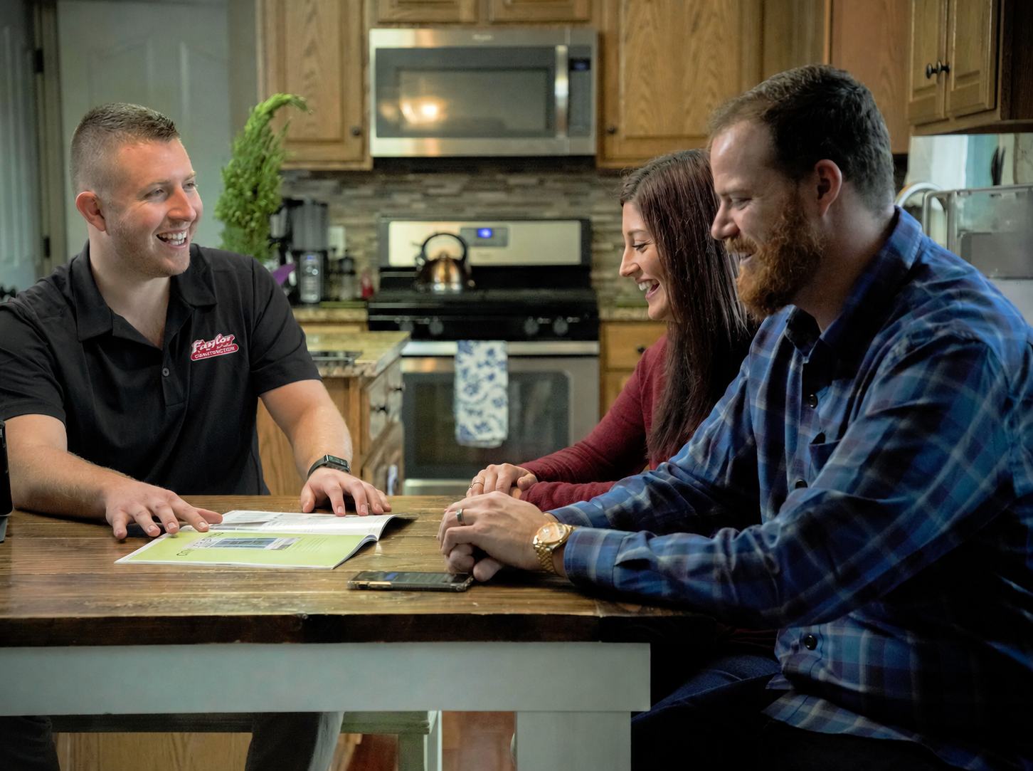 Four people sitting around a kitchen table, smiling and talking, with an open magazine, phone, and a pen on the table.