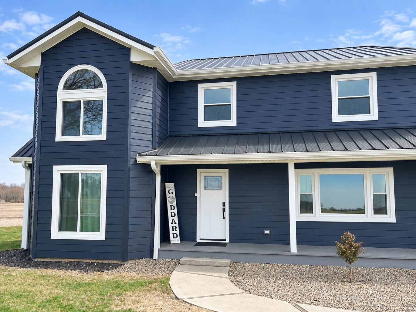 A two-story blue house with white trim, large windows, and a metal roof, with a small front porch, a curved walkway, and a sign that reads 'G DARD' leaning against the house.