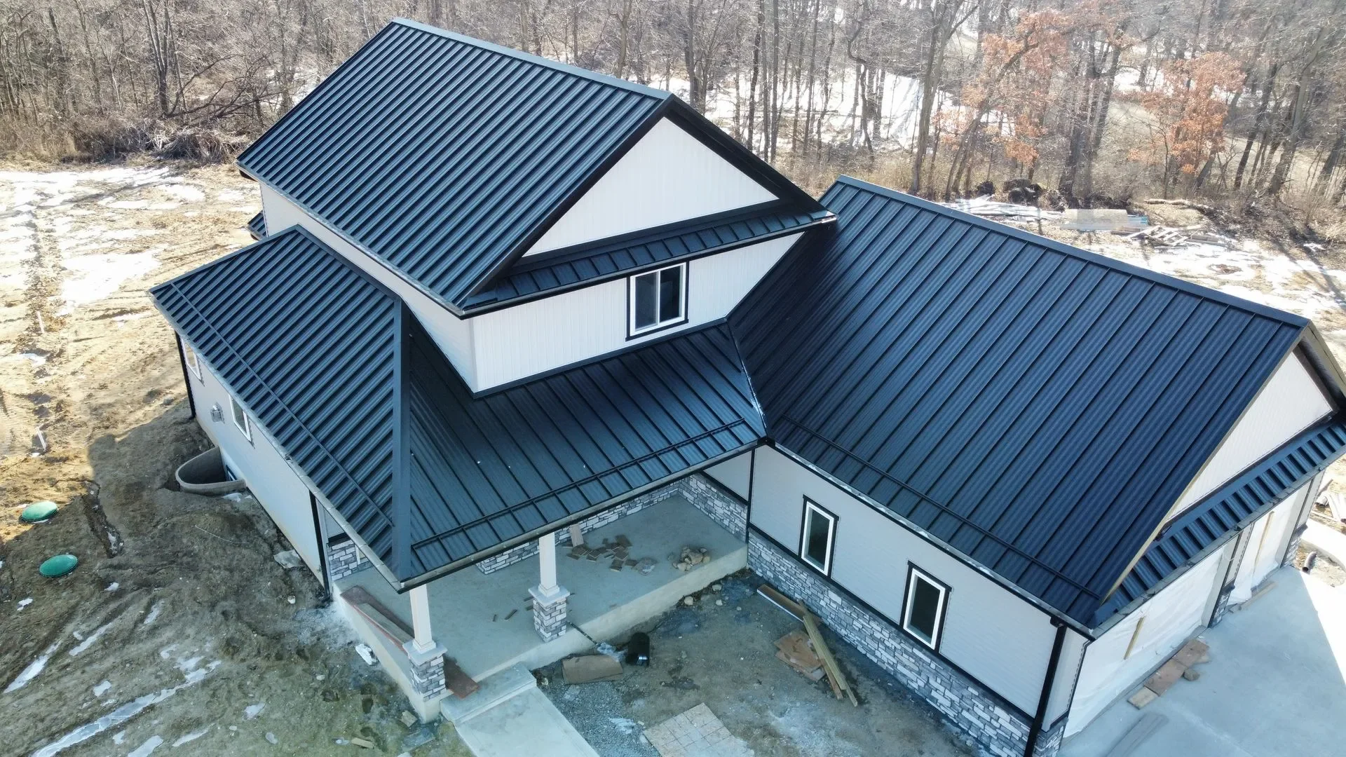A house under construction with black metal roofing, white siding with black trim, and stone accents at the base. The house has a covered porch and multiple roof sections, surrounded by a dirt yard and leafless trees in the background.