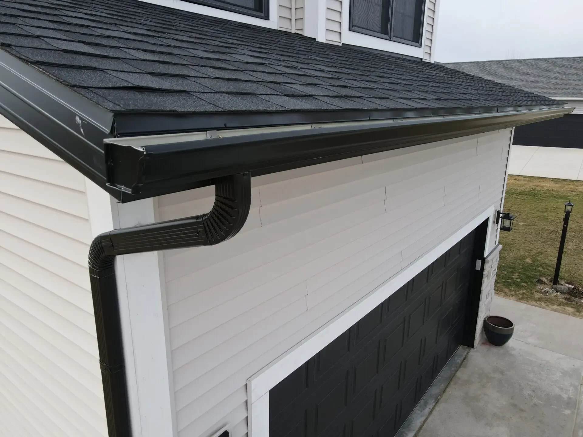 A house with contrasting white and black siding, featuring a black roof with asphalt shingles, black gutters, and a black garage door. There is an outdoor lamp post and a small planter on the patio.