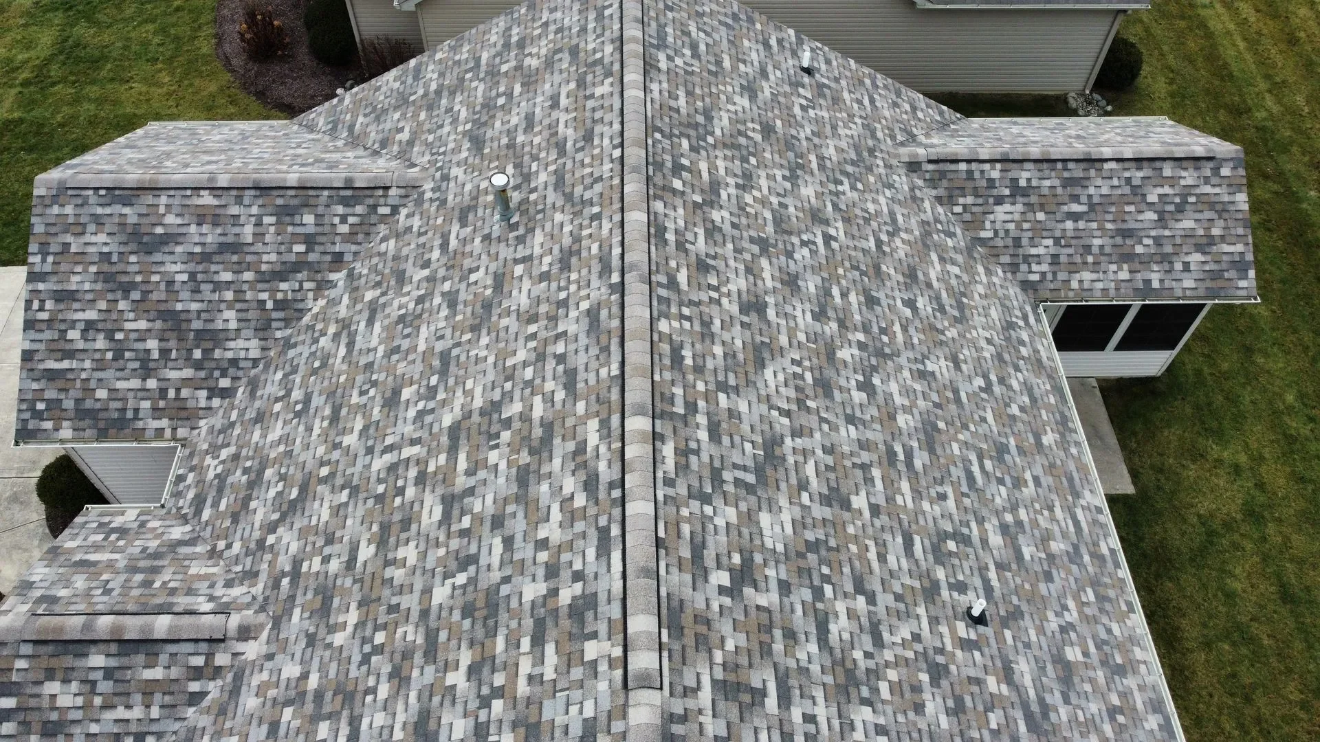 Aerial view of a house roof with multi-colored shingles, central ridge, and vents, surrounded by grass and neighboring houses.