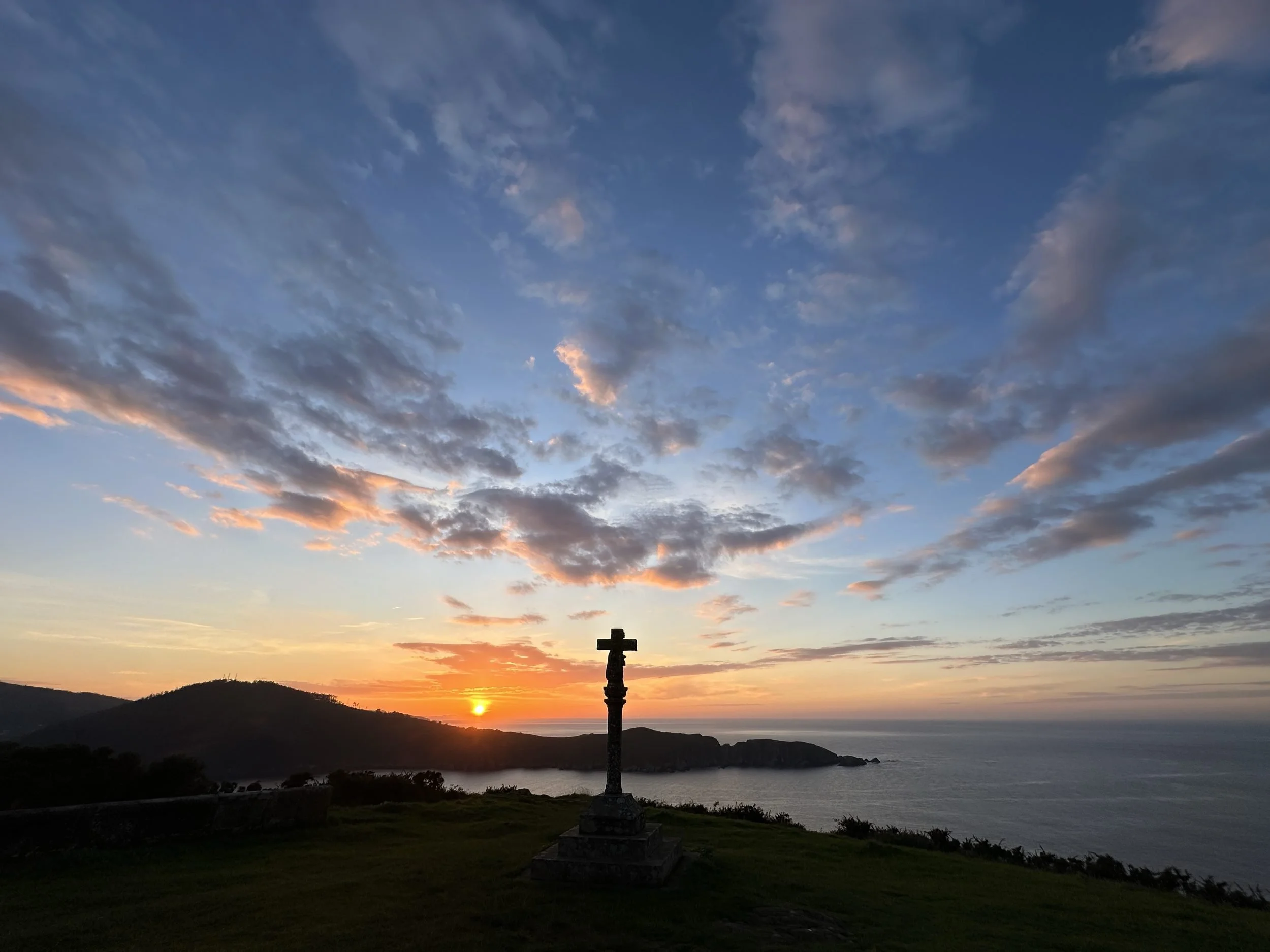 Panoramic view featuring the silhouette of a traditional Galician stone cross (cruceiro) on a grassy hill overlooking a golden ocean sunset and dramatic headlands, photographed by Seña Studio.