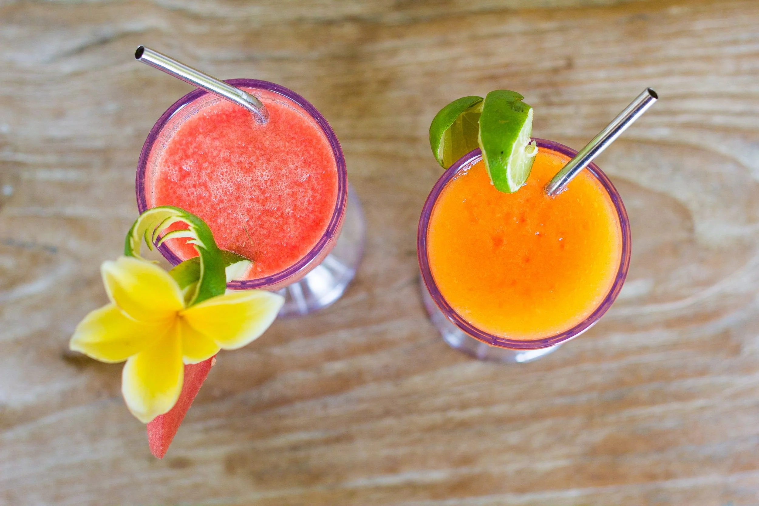 Directly overhead shot of two chilled fruit smoothies with metal straws—one pink watermelon garnished with a frangipani flower and one orange papaya garnished with lime—on a rustic wooden table, photographed by Seña Studio.