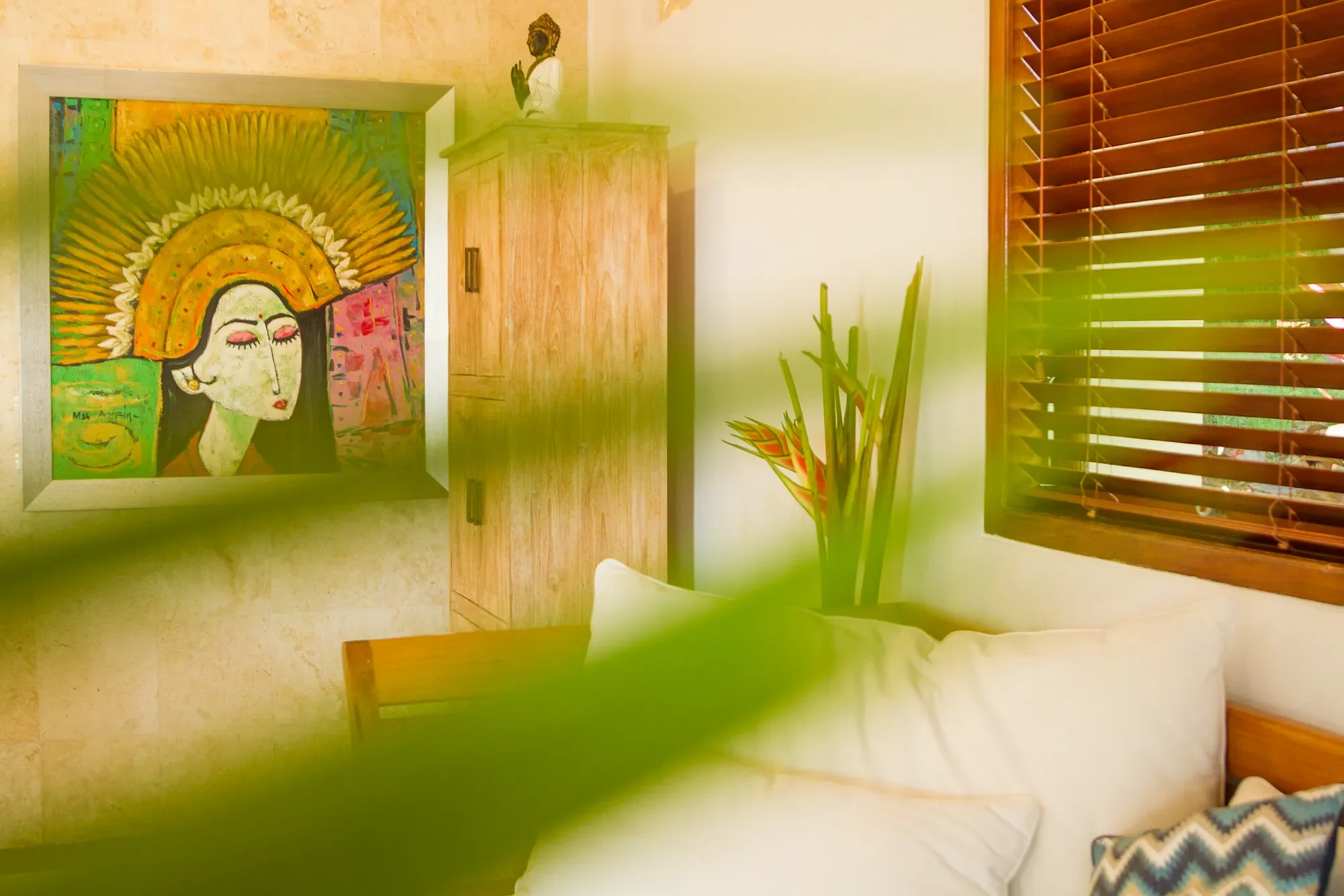 Interior detail of a living area at Aquamarine Beach Villas, Bali, featuring a large colorful Balinese dancer painting, wooden furniture, a Buddha statue, and tropical plants, photographed by Seña Studio.