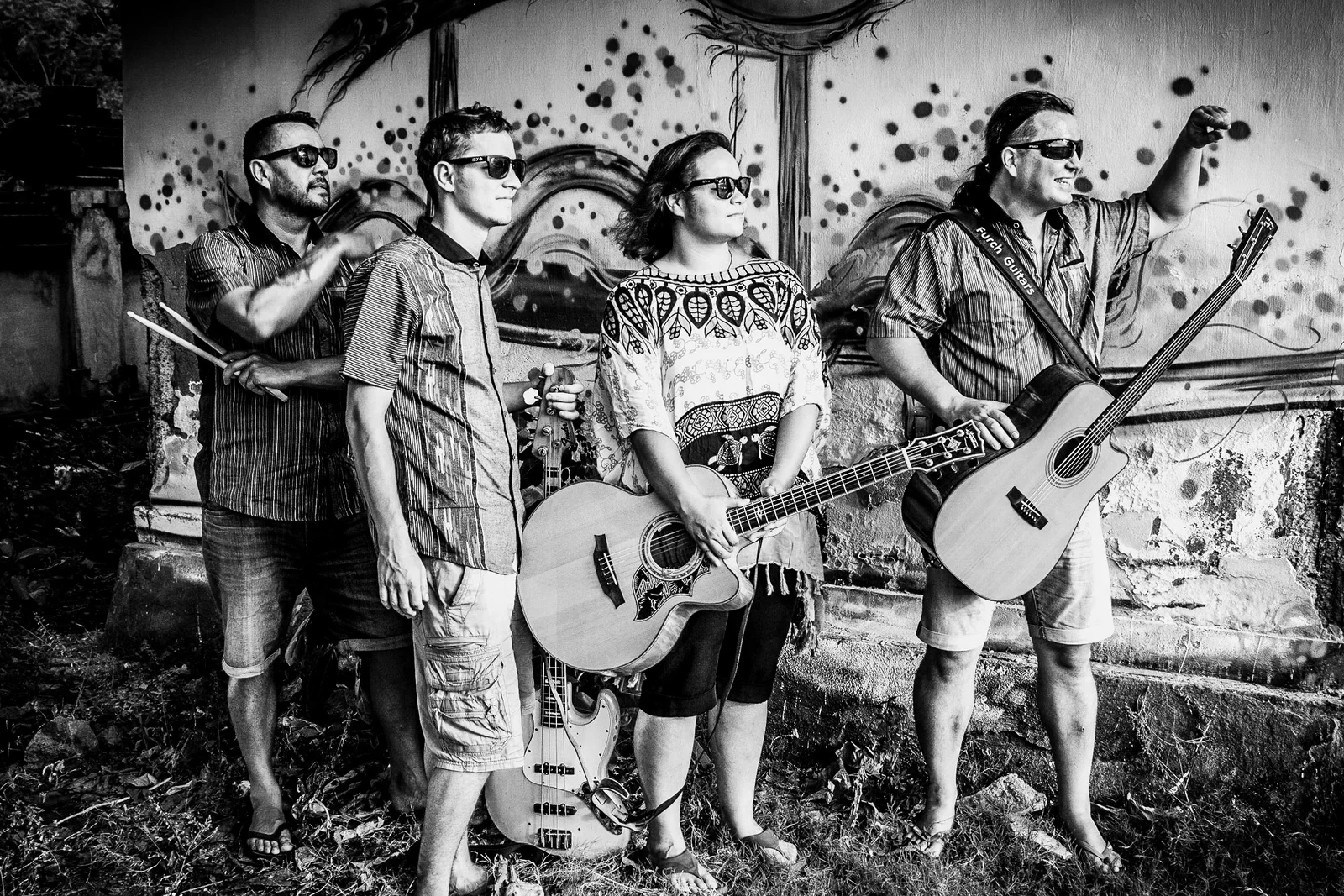 Gritty black-and-white group portrait of a four-piece band - led by musician Lucy Dobrovodska - holding guitars and drumsticks while wearing sunglasses in front of a painted graffiti wall, photographed by Seña Studio.