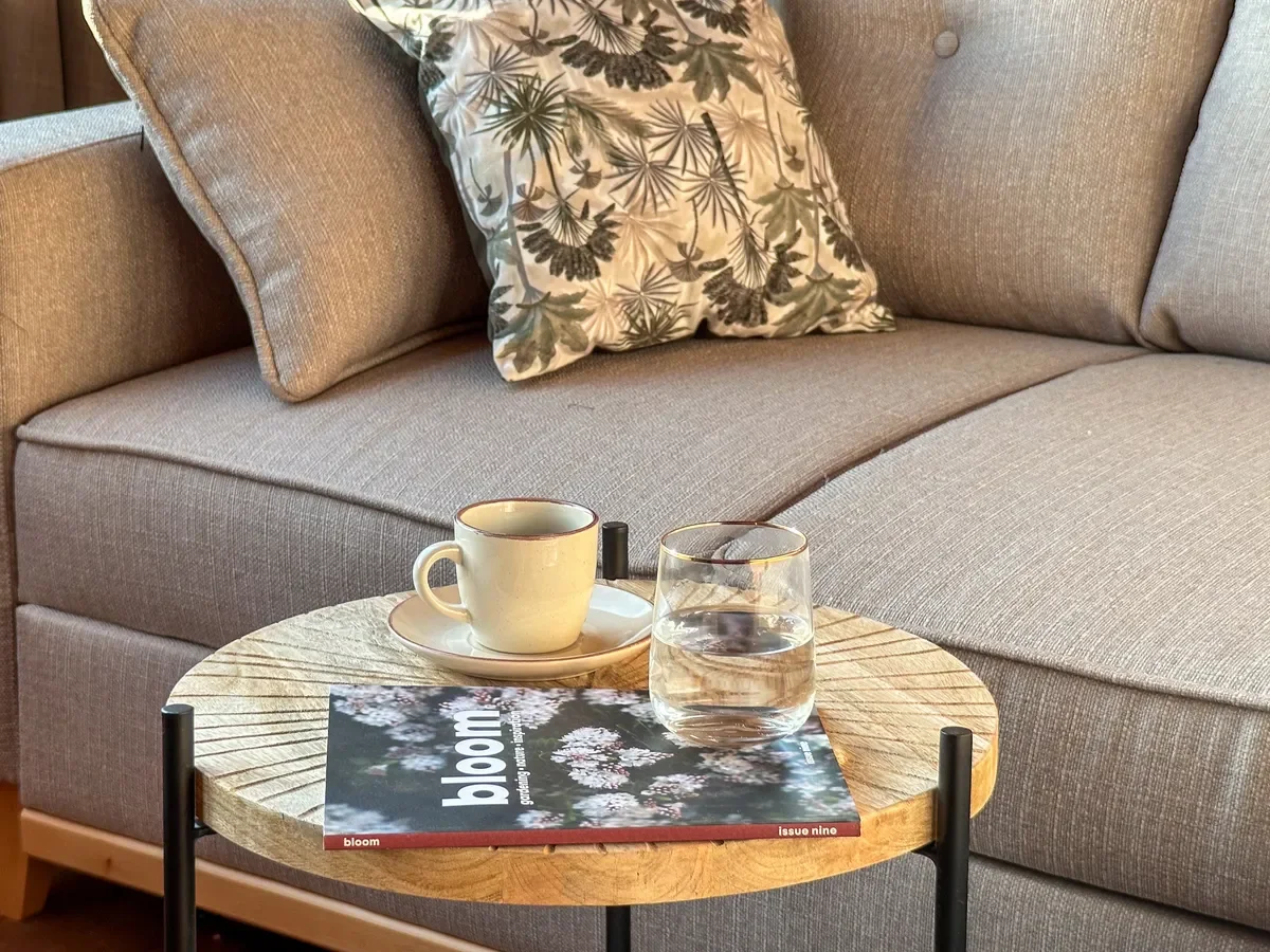 Cozy interior detail featuring a wooden side table with a coffee cup, glass of water, and magazine next to a modern grey sofa, photographed by Seña Studio.