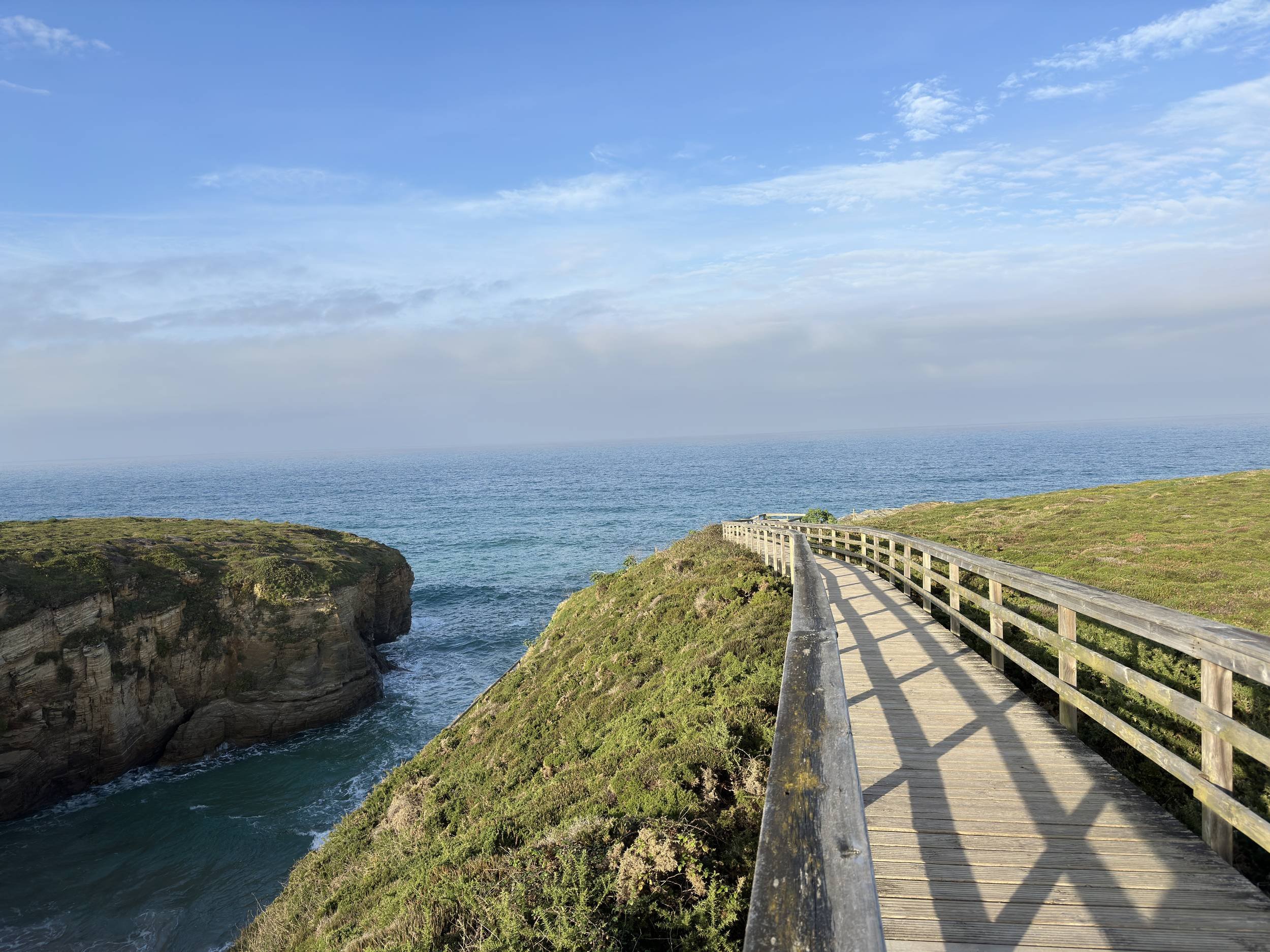 Wooden boardwalk trail winding along grassy cliff tops leading toward open Atlantic Ocean views and sea inlet caves in Northern Galicia, photographed by Seña Studio.