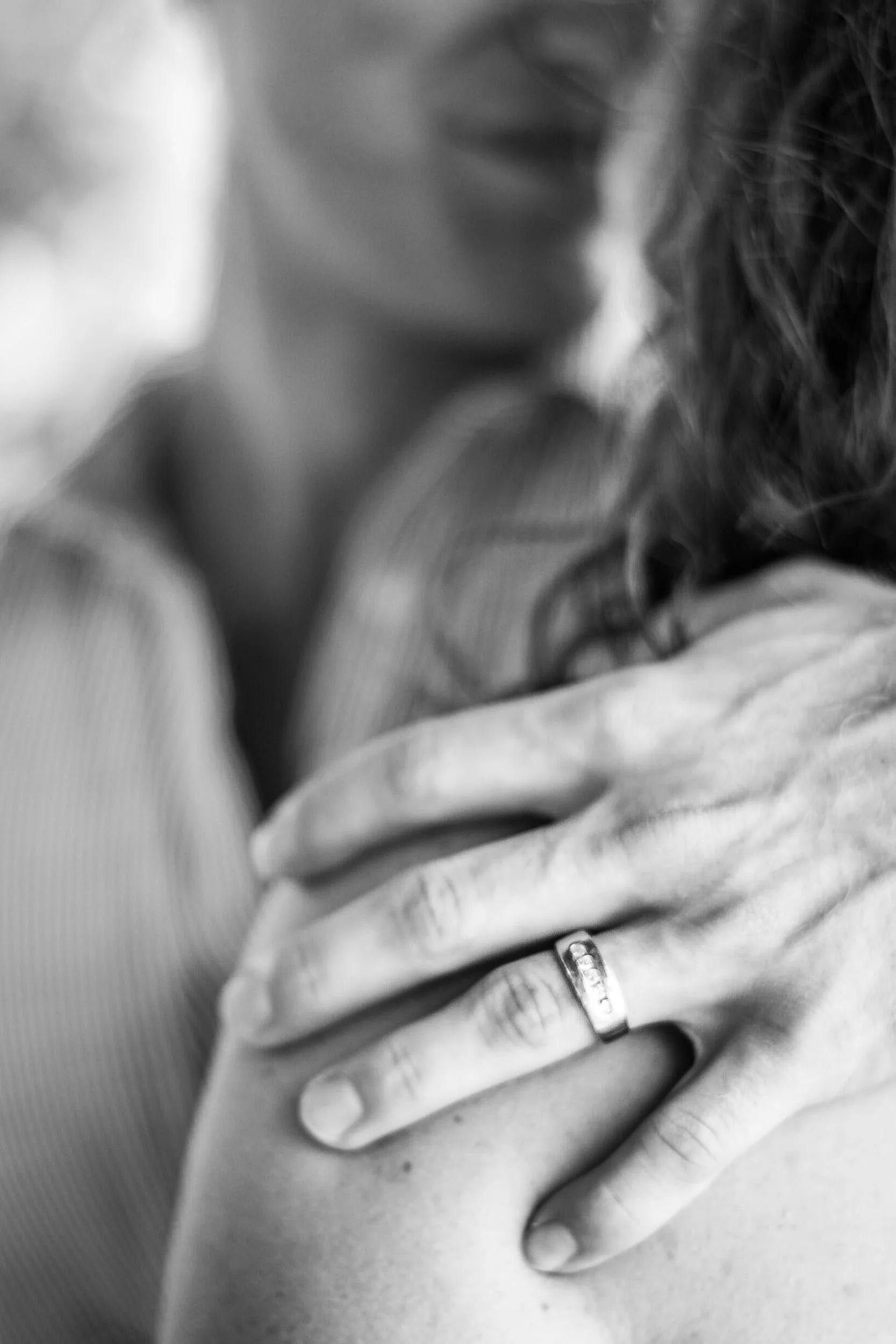 Intimate black-and-white macro portrait of a man's hand resting tenderly on a woman's shoulder during a close embrace, highlighting his silver wedding ring, photographed by Seña Studio.