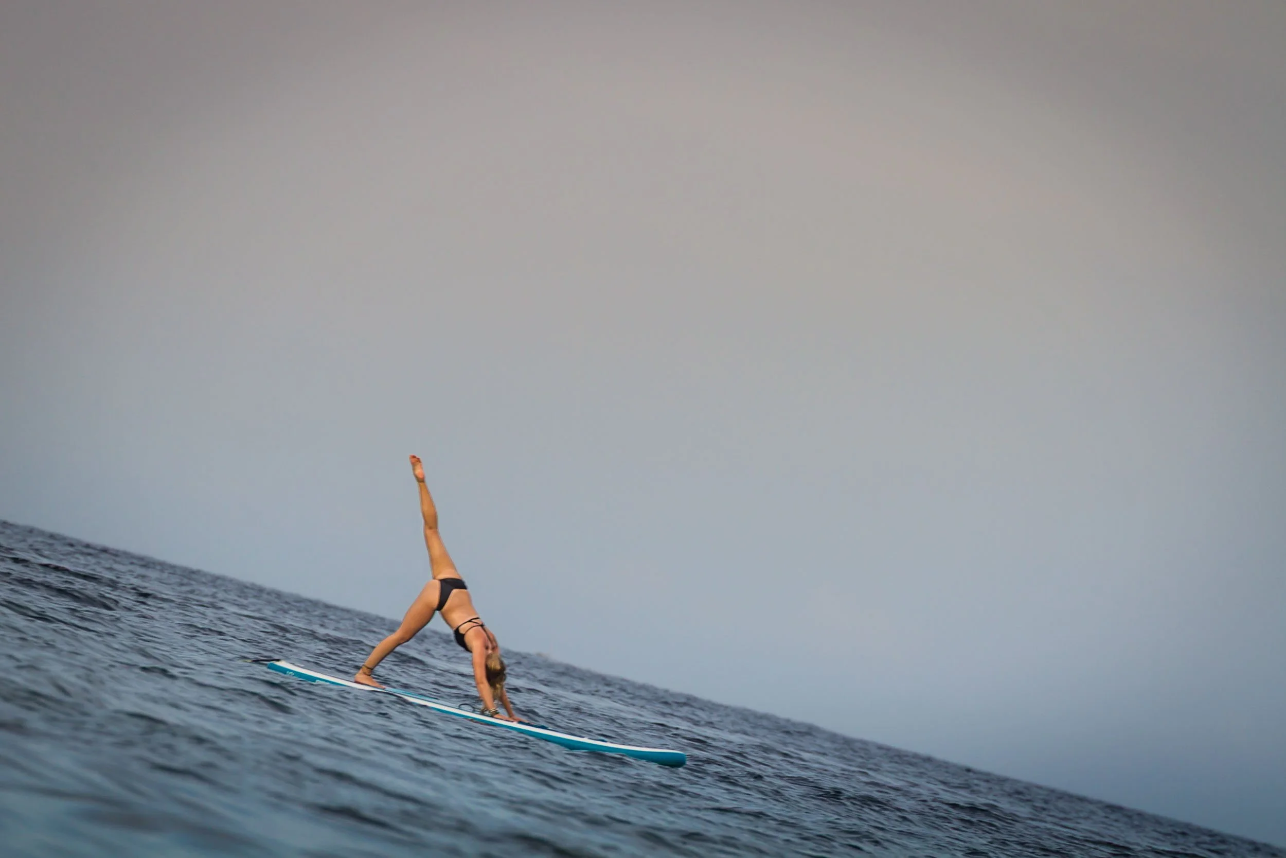 Wide-angle shot of a woman practicing a three-legged downward dog pose on a paddleboard floating on open ocean waters, photographed by Seña Studio.