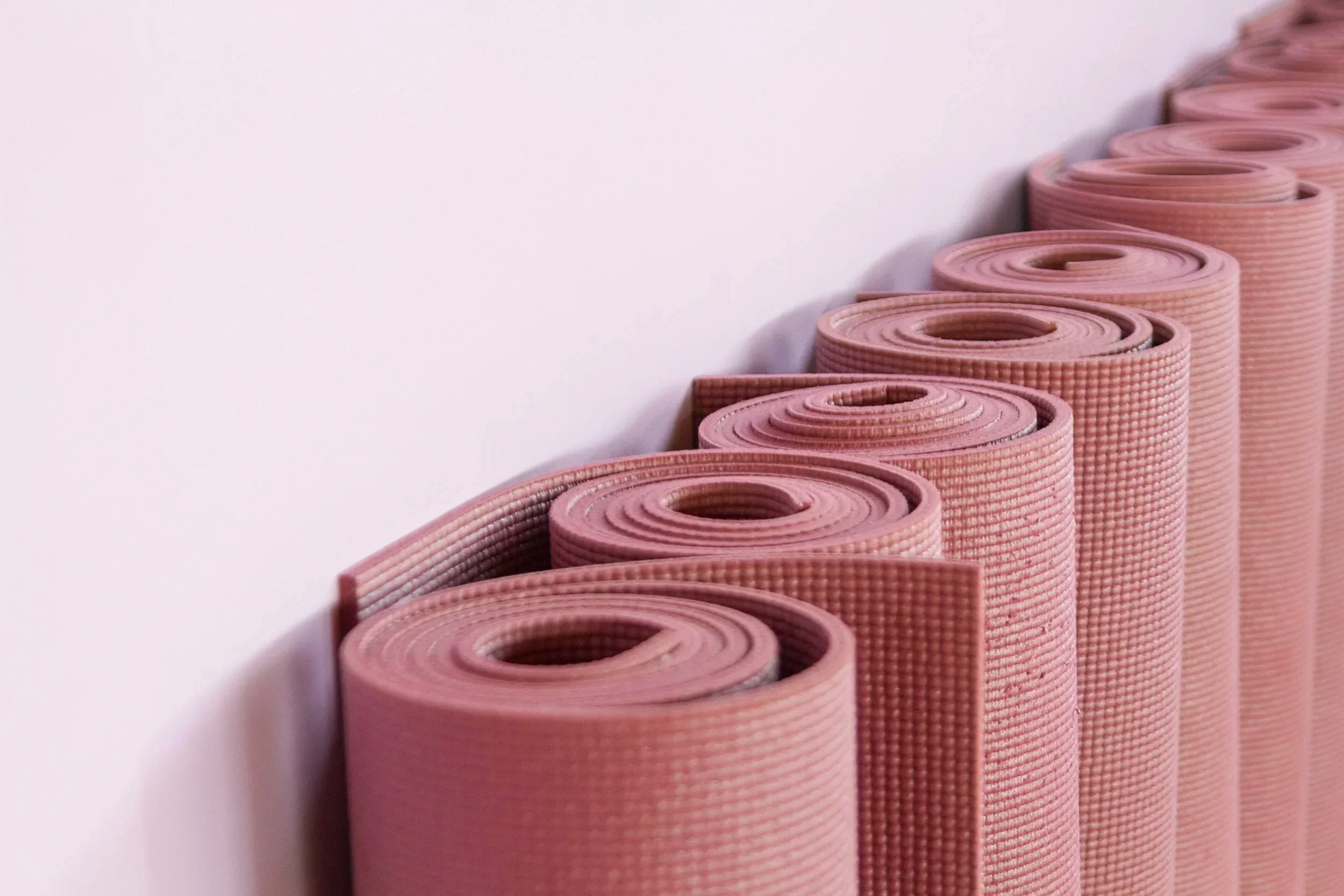 Minimalist close-up photograph showing a neat diagonal row of rolled dust-pink yoga mats resting against a light wall in a wellness studio space.