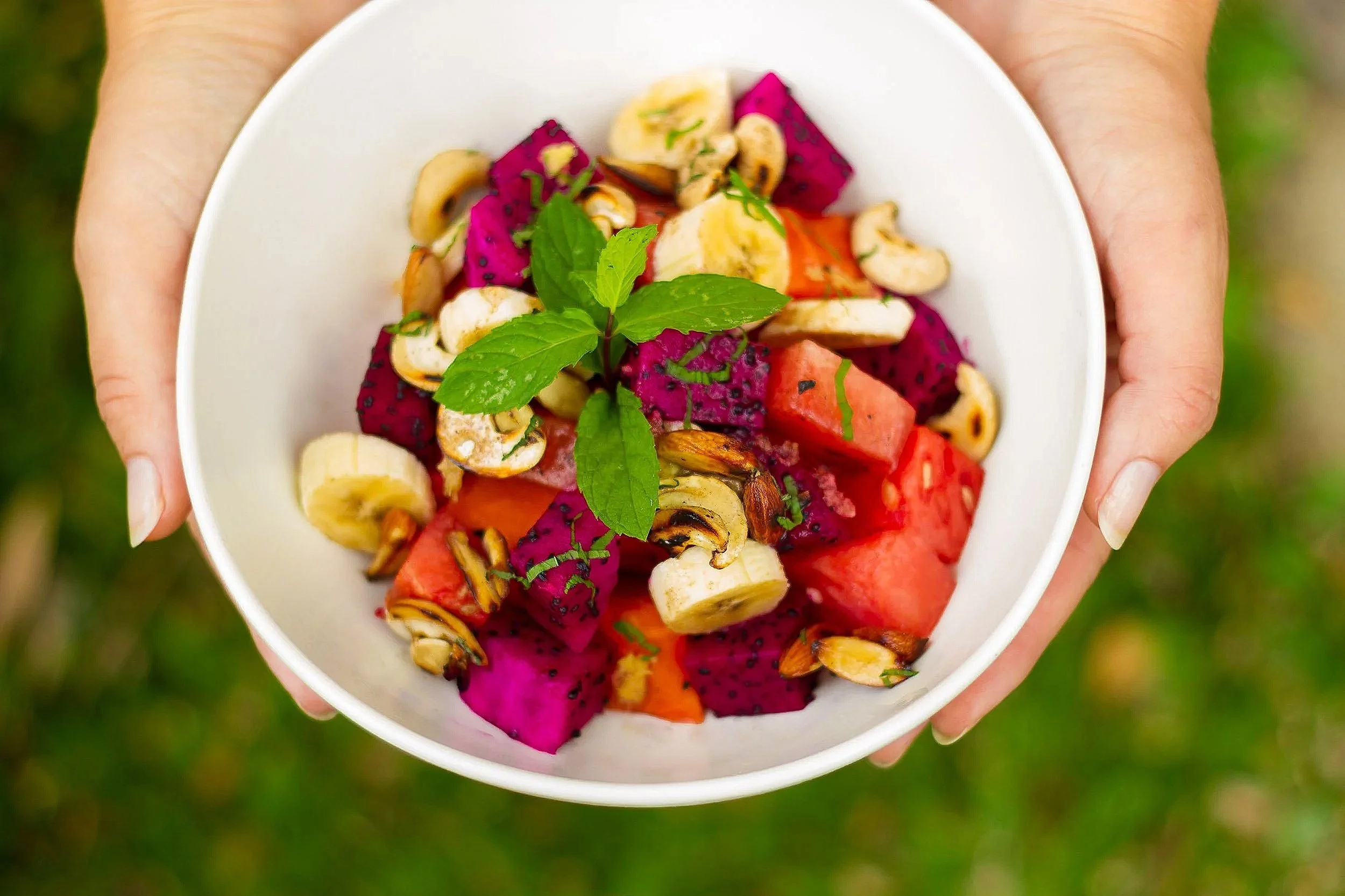 A top-down view of two hands holding a white bowl filled with fresh diced dragon fruit, watermelon, sliced bananas, toasted cashews, and fresh mint leaves.