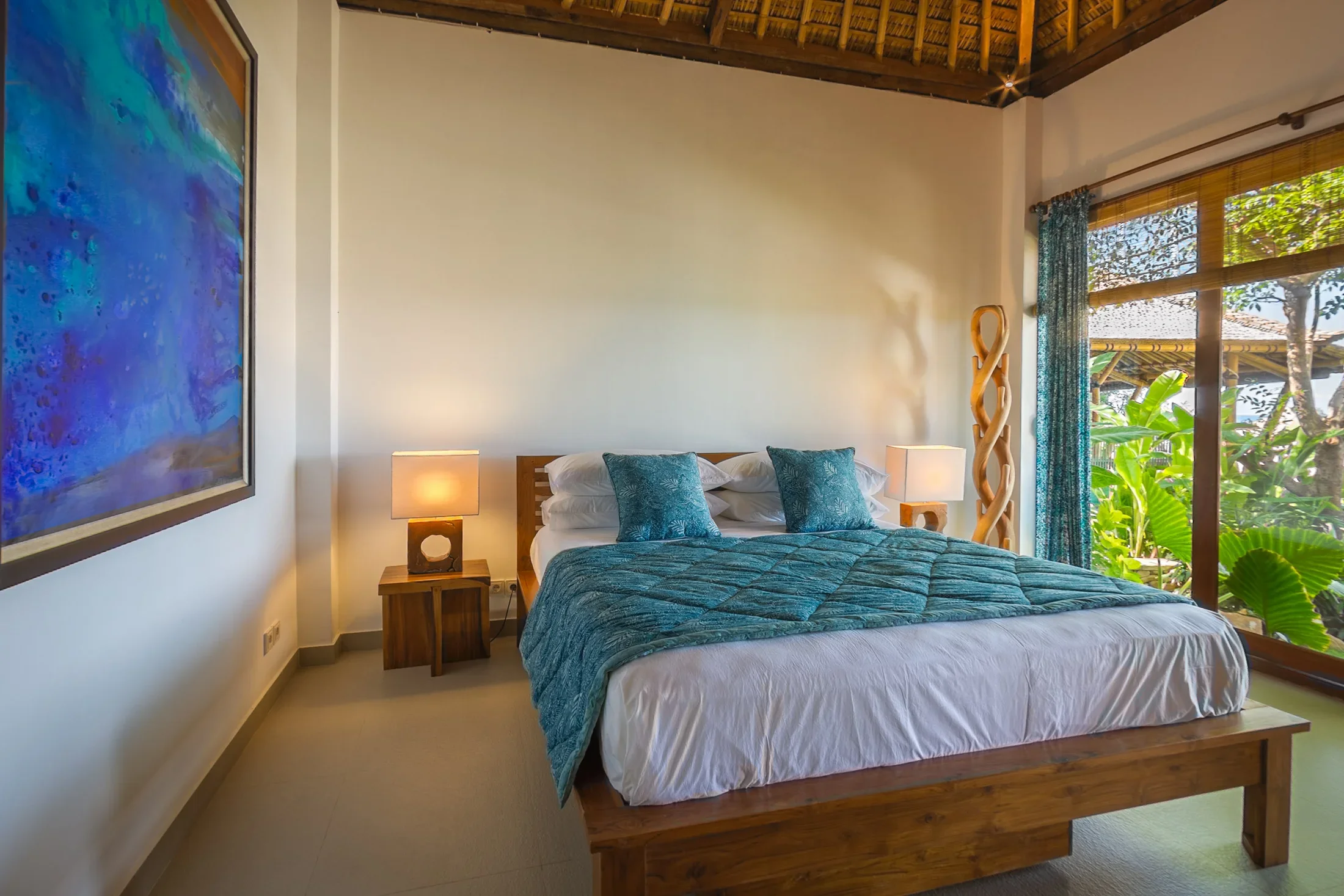 The interior of a contemporary boutique hotel room, featuring a large wooden bed with crisp white linens and teal blue patterned pillows and duvet. An enormous, vibrant abstract blue and purple painting hangs on the left wall. To the right, floor-to-