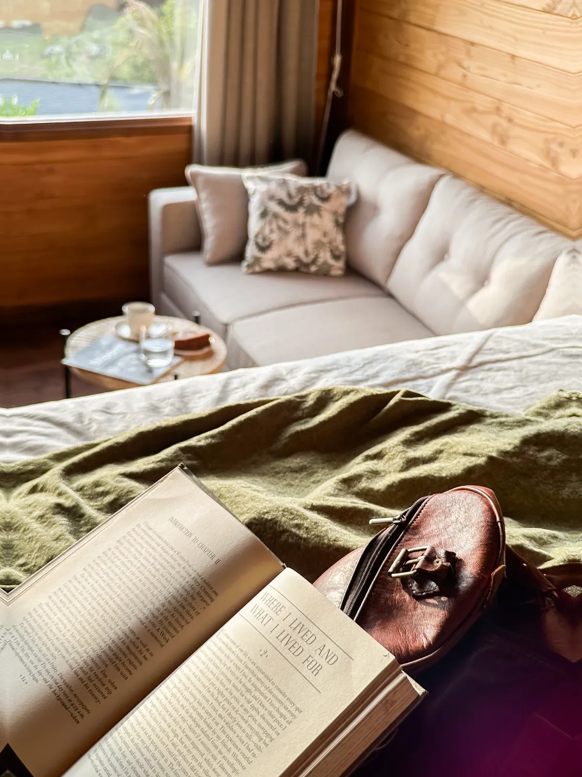 Indoor scene of a cozy living room with a white couch with patterned cushions, a wooden wall, and a window with curtains. The view from the bed shows a nightstand with a glass of water and a small tray. A book, a brown leather bag, and a green blanket are in the foreground.