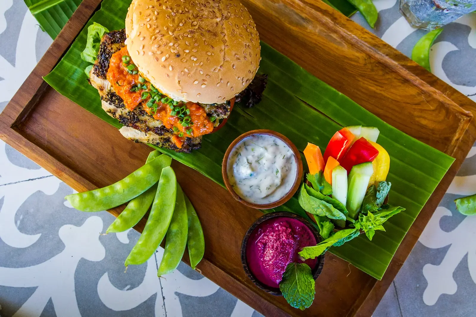 Overhead view of a gourmet chicken burger served on a wooden platter with fresh vegetable sticks, snap peas, and dipping sauces at The Griya Villas & Spa in Amed, Bali, photographed by Seña Studio.