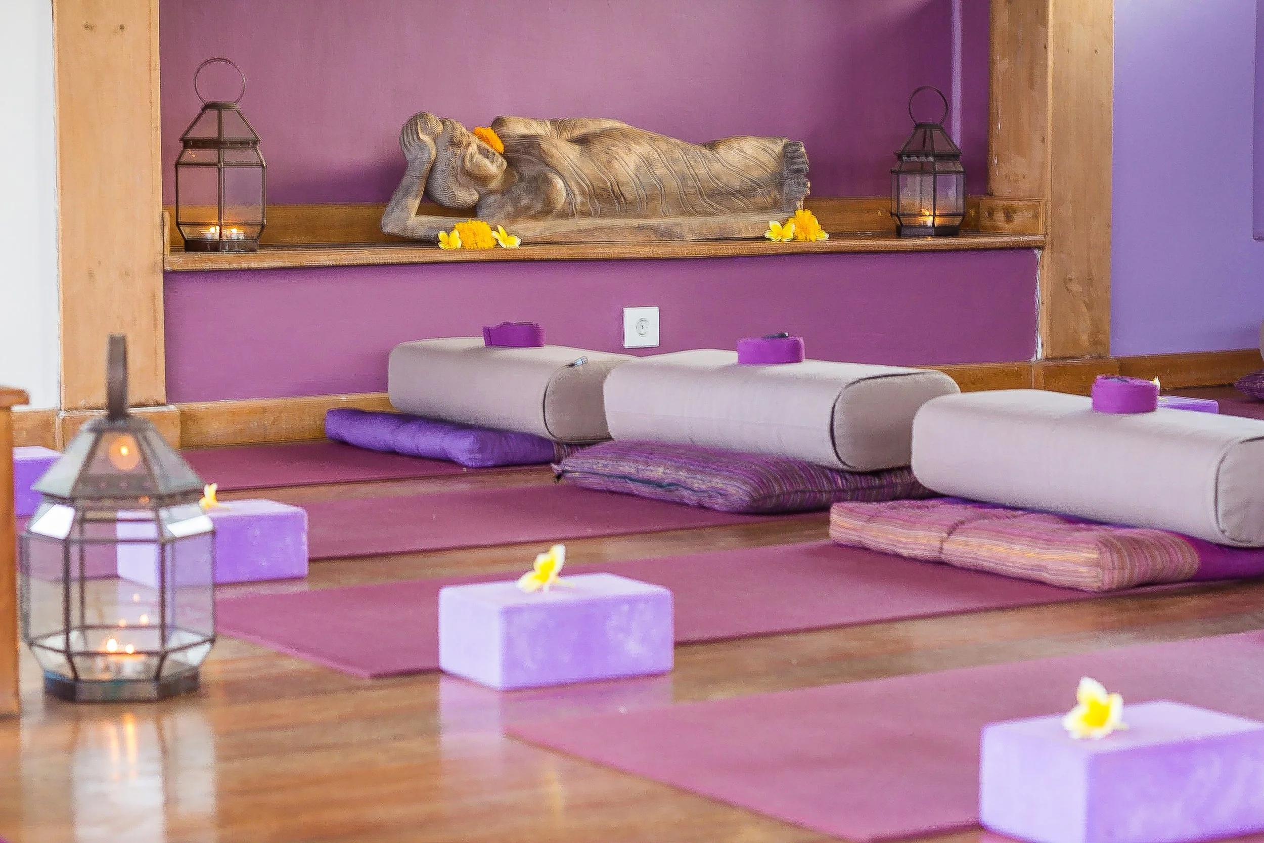 Interior view of a serene yoga studio with purple mats, bolsters, foam blocks adorned with frangipani blossoms, and a carved wooden reclining Buddha statue on a shelf, photographed by Seña Studio.