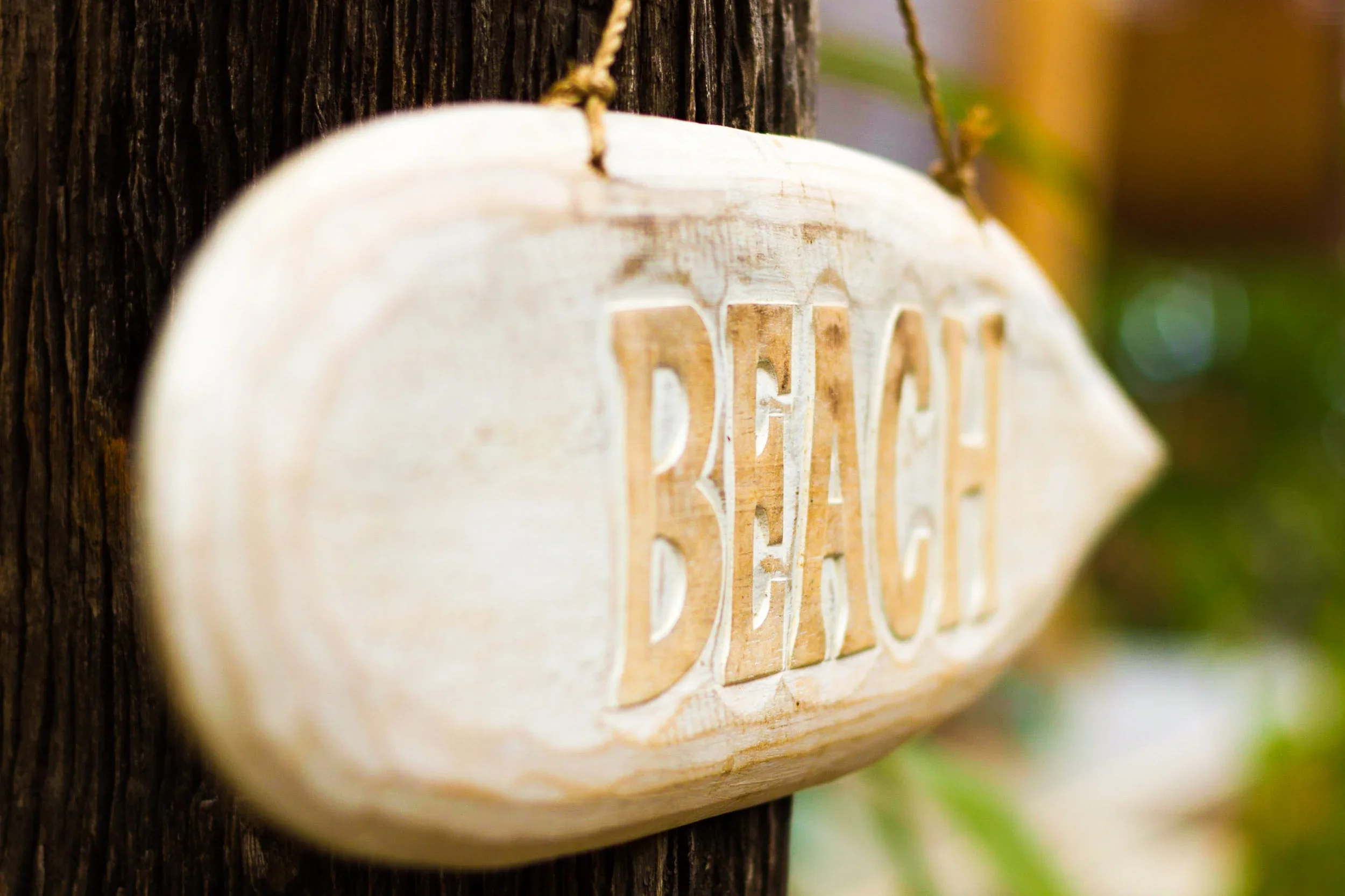 A very close-up, shallow depth-of-field photograph of a rustic, hand-carved and whitewashed wooden sign. The word "BEACH" is visible, carved into the wood. The sign hangs by a natural twine loop from a textured, dark wooden post.