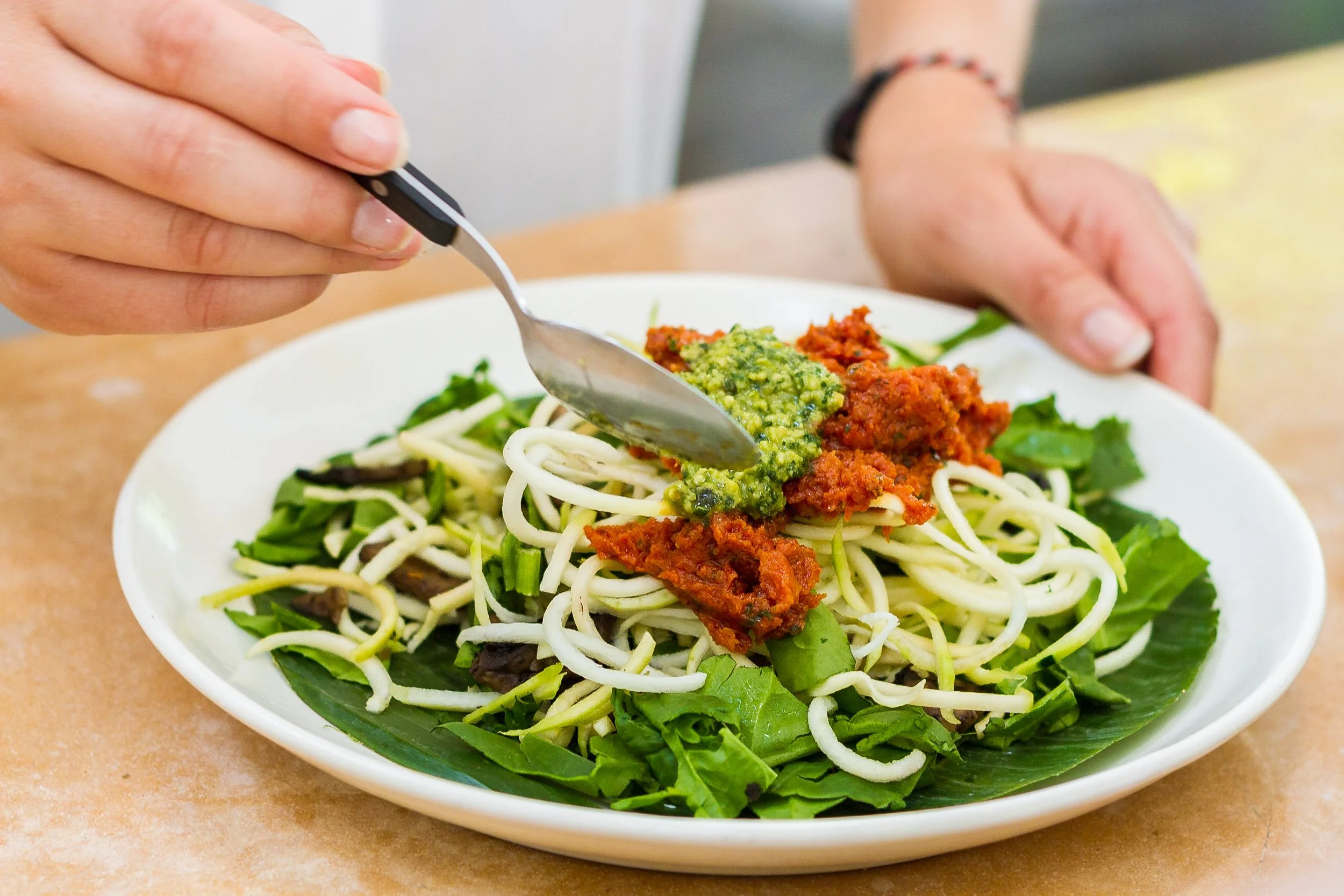 A close-up of a spoon adding green pesto to a raw zucchini noodle salad topped with rich sun-dried tomato red sauce on a white plate.