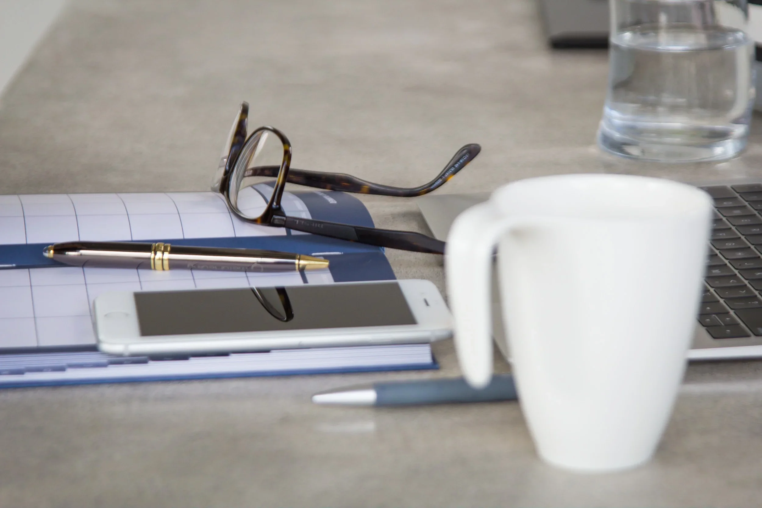 Still life of a modern workspace setup featuring an open planner, eyeglasses, pen, smartphone, laptop, and a white coffee mug on a grey desk.