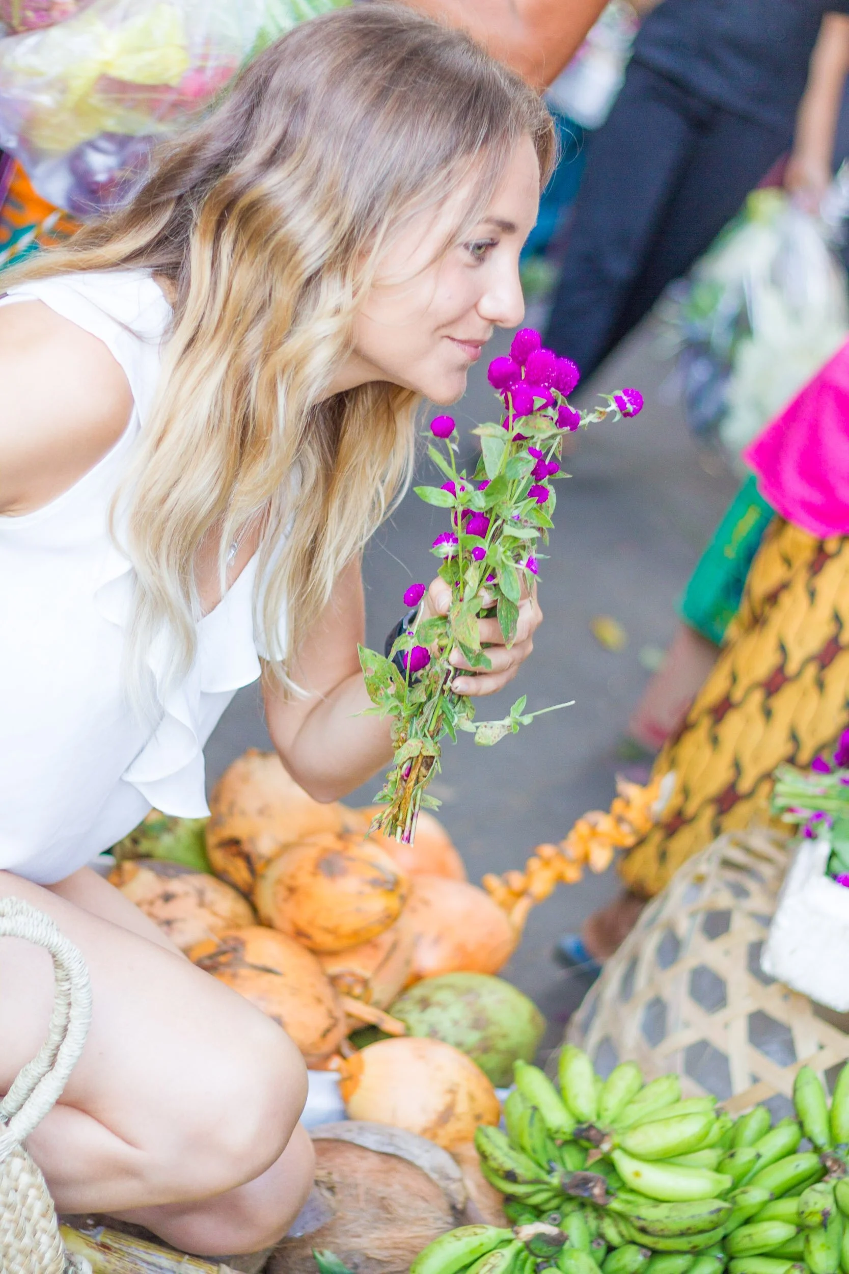 A smiling woman gently smells a small bouquet of vibrant purple globe amaranth flowers at a local market stand. Ubud, Bali.