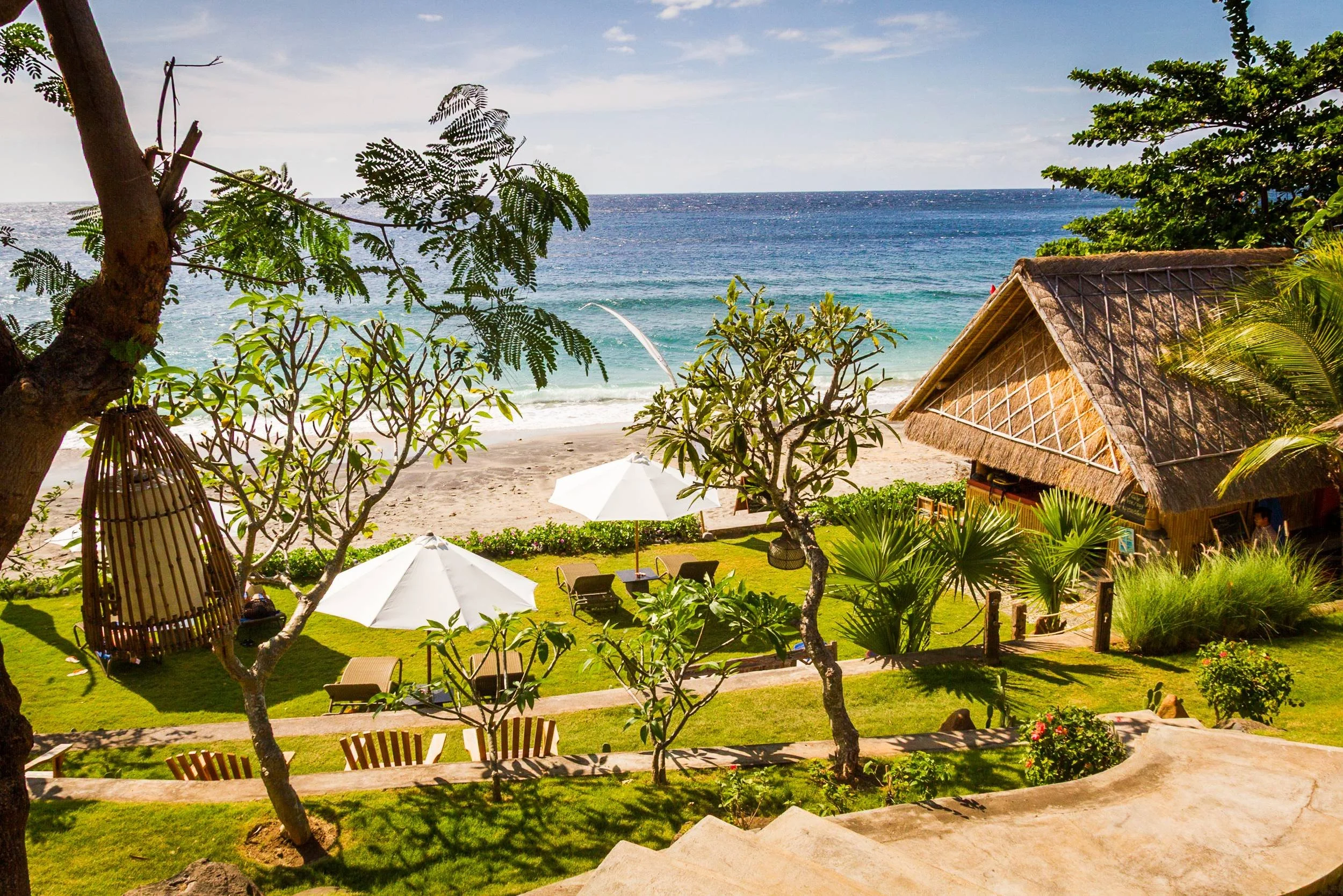 Elevated view of a tropical beachfront garden featuring white sun umbrellas, a thatched bamboo bar, and ocean views at Aquamarine Beach Villas in Amed, Bali, photographed by Seña Studio.