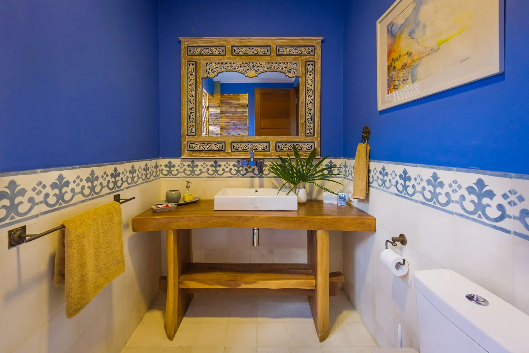 Bright resort bathroom interior featuring vivid blue walls, decorative patterned tiles, a custom wooden vanity with a white vessel sink, and a carved mirror frame.