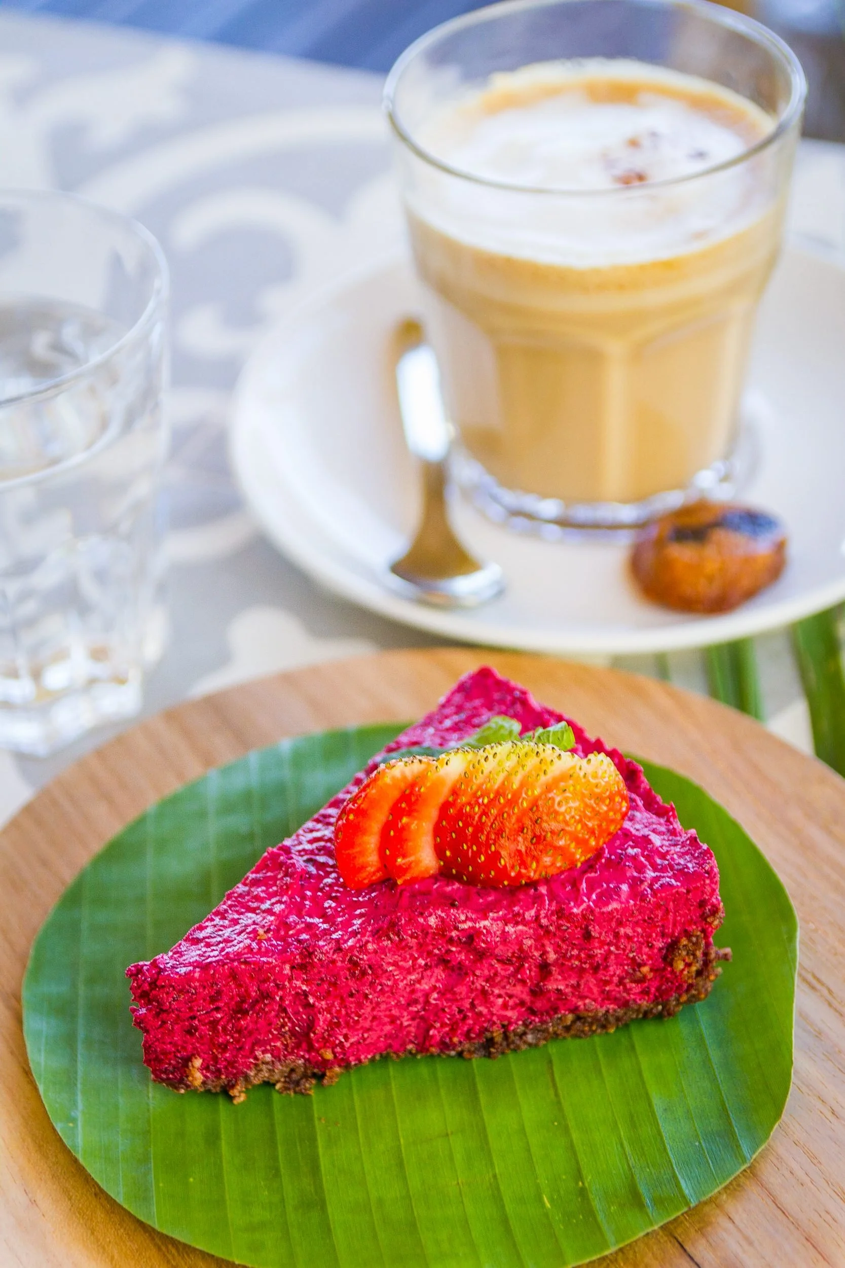 A slice of bright magenta raw dragon fruit cheesecake garnished with fresh strawberries, served on a banana leaf with an iced coffee drink in the background, photographed by Seña Studio.