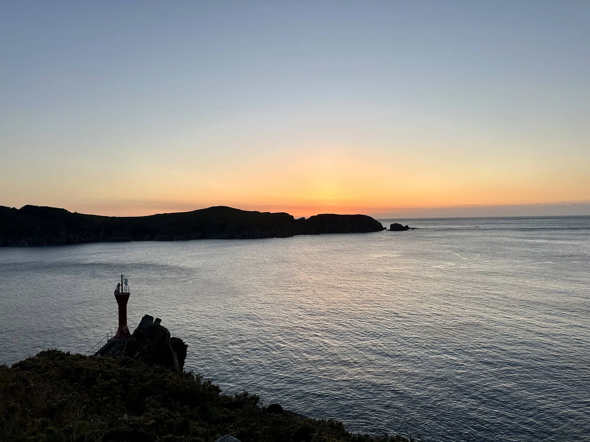 Golden sunset glowing over a calm sea horizon with the silhouette of a red coastal navigational beacon on rocky cliffs in Northern Galicia, photographed by Seña Studio.