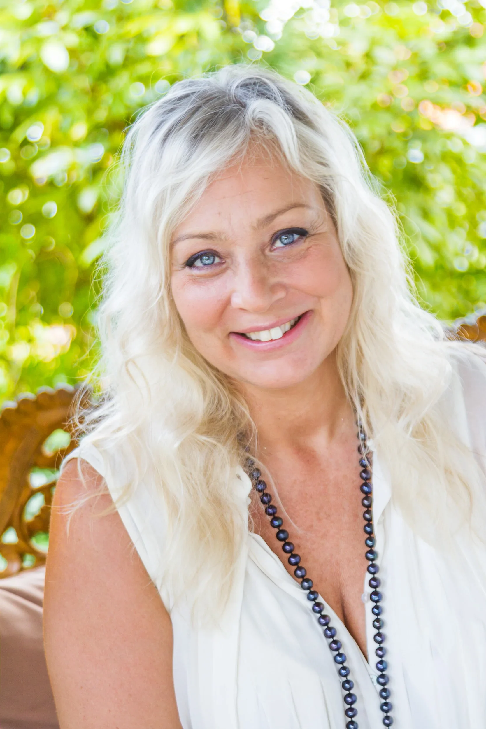 Close-up portrait of a smiling blonde woman with blue eyes wearing a white top and a long dark pearl necklace.