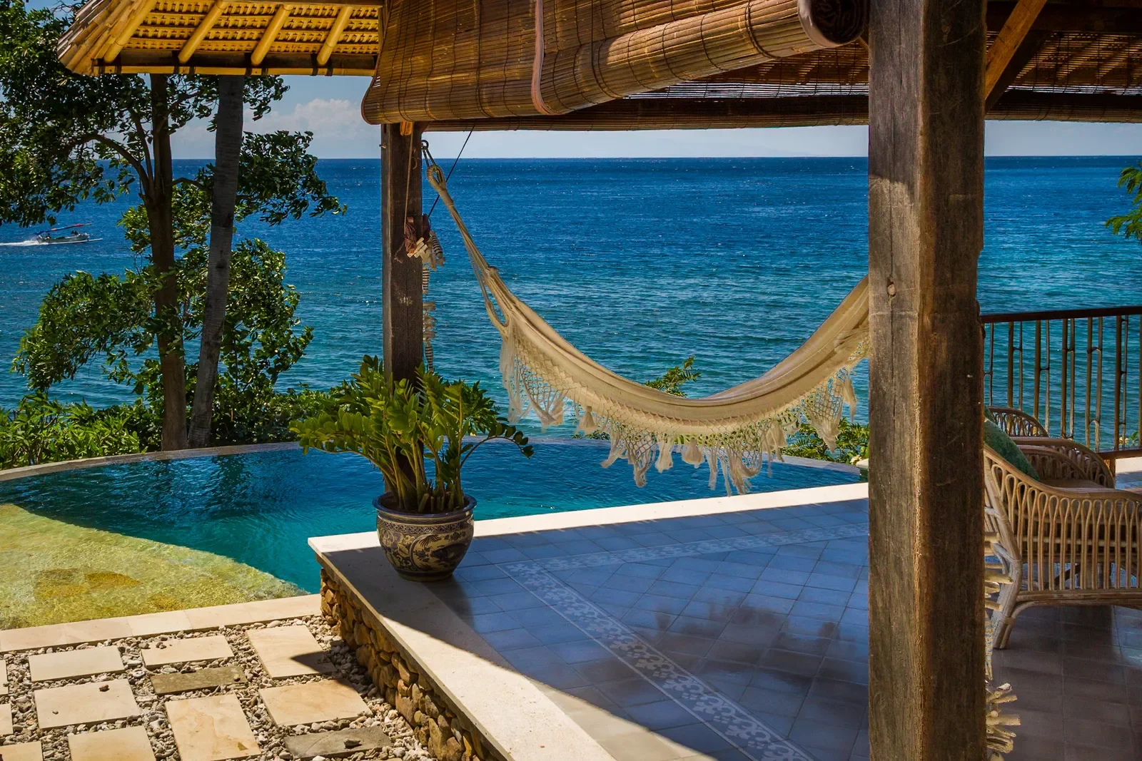 A relaxing view from a covered open-air terrace, featuring a cream-colored woven hammock with fringe hanging between rustic wooden posts, next to a private infinity pool and an ornate blue and white ceramic planter, overlooking the ocean under a clea